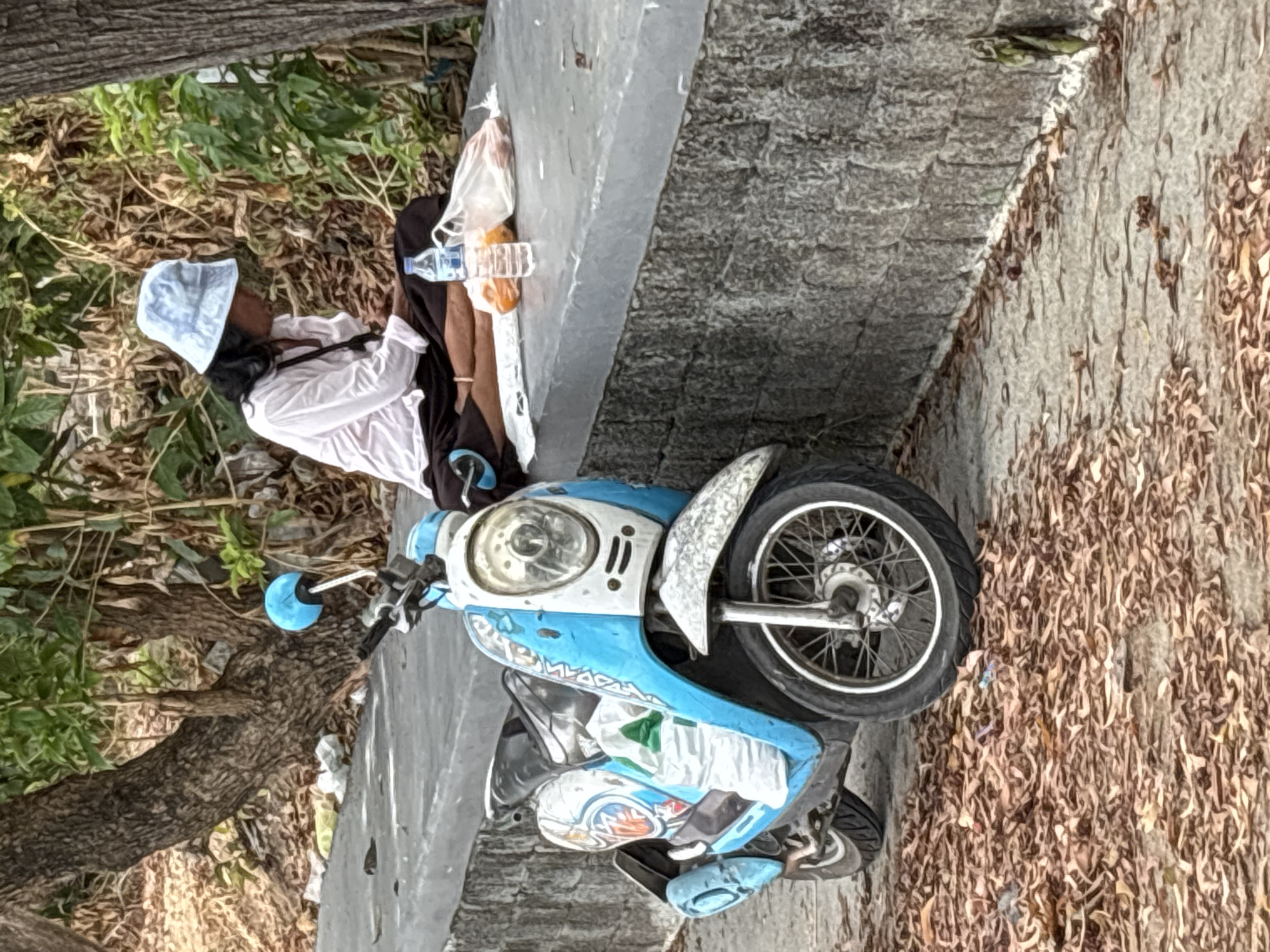 A person wearing a white shirt and light blue hat is sitting with his back to a low concrete wall in the city of บ้านบางรักษ์, Thailand. A blue and white motor scooter is parked adjacent to the wall, with its front tire resting near a pile of dried leaves. Water bottles and a plastic bag are positioned near the person on the wall. The area also contains trees, bushes, and a grey pavement surface.