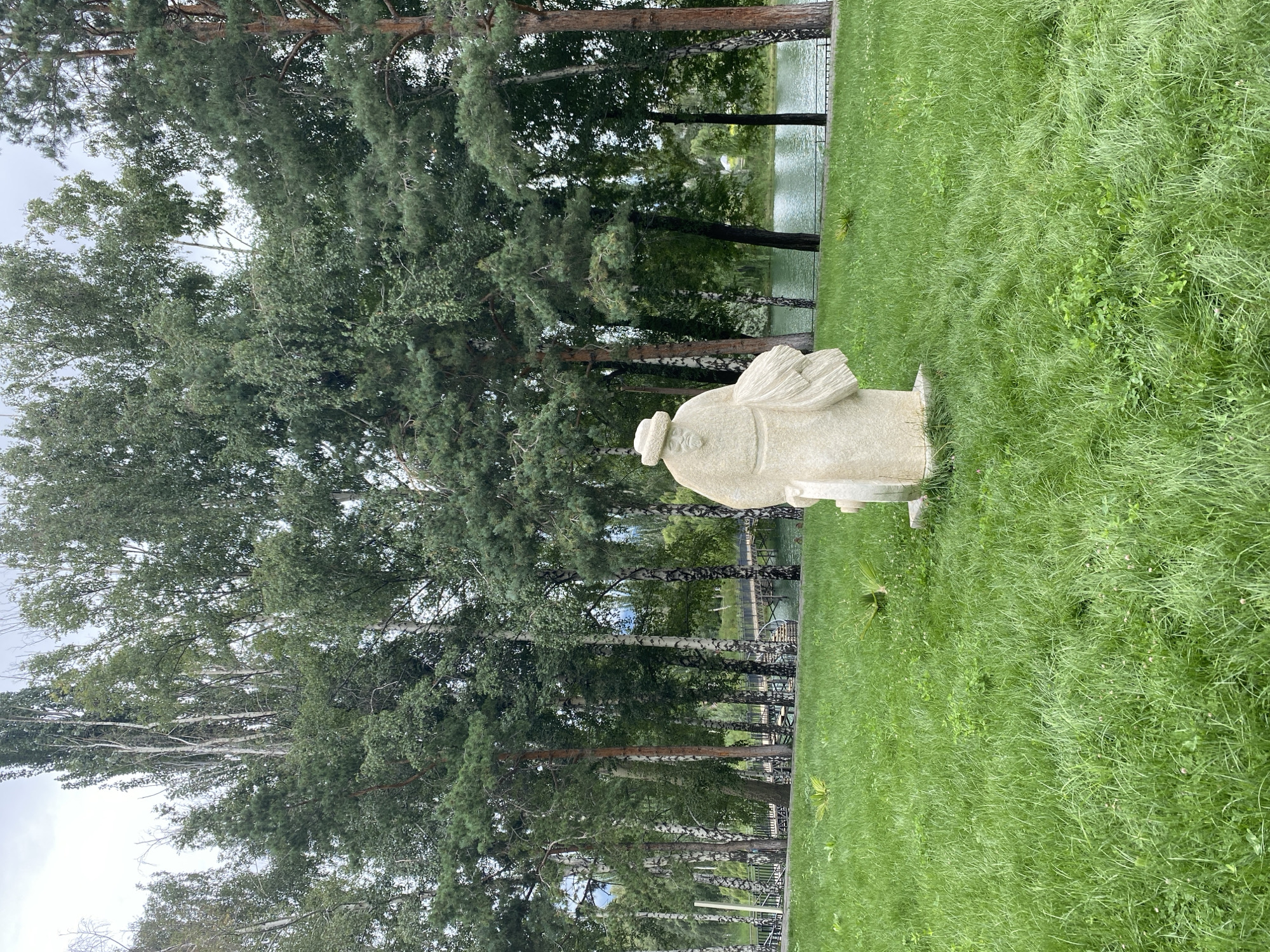 A stone sculpture of a man wearing a hat and traditional clothing stands in a grassy park. The sculpture is positioned in the foreground, to the right of the center. Behind the sculpture, a body of water is visible, with trees lining its banks. A bridge can be seen spanning the water in the distance. The sky is overcast. The location is the park in Cholpon-Ata, Kyrgyzstan.