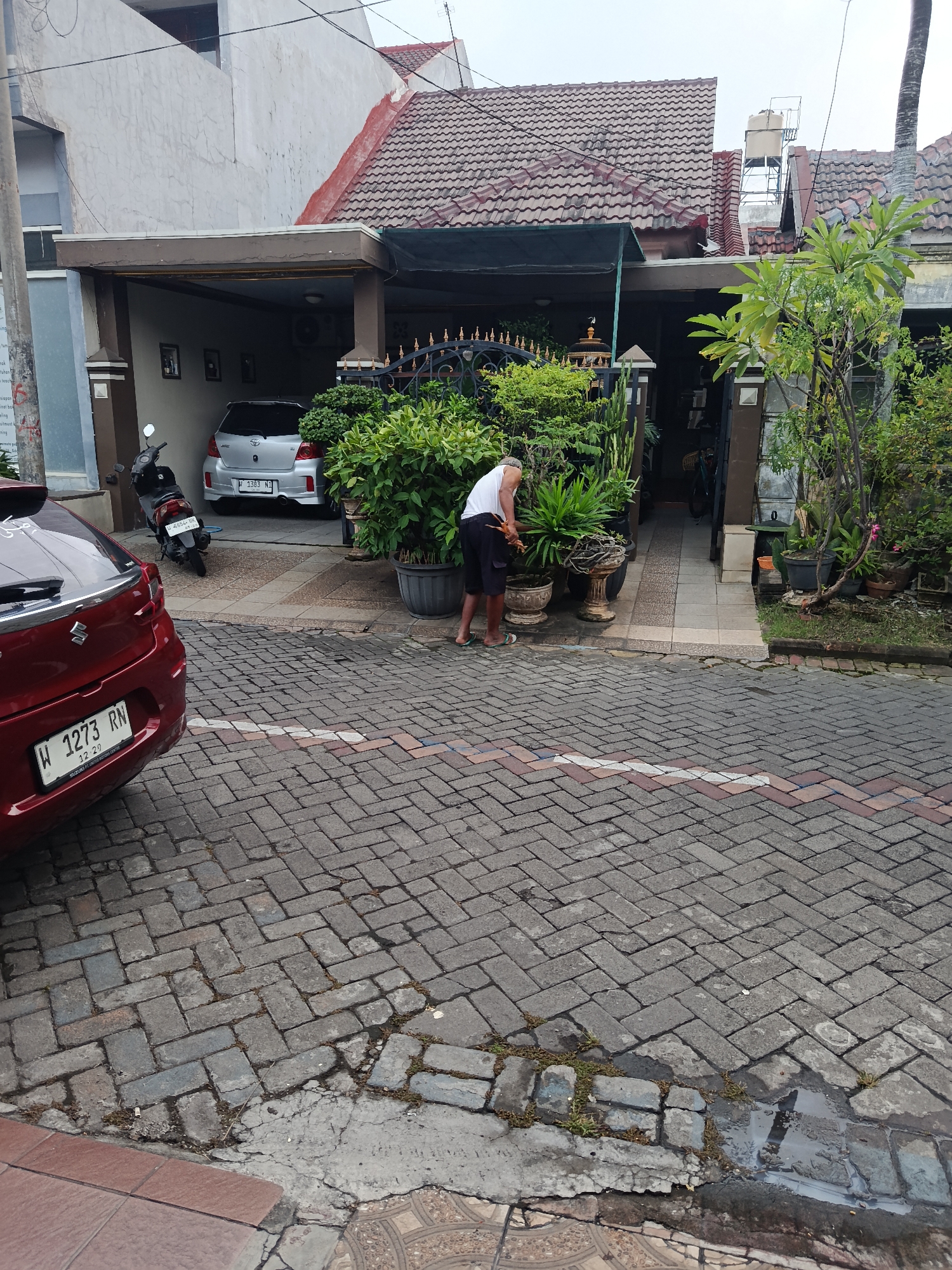 The image depicts a residential street in Keboananom, Indonesia, with a focus on the front of a house. A man, wearing a white t-shirt and dark shorts, is tending to plants in pots in front of the house. He appears to be using a tool to cultivate the plants. The house has a tiled roof and an open carport where a silver car is parked, alongside a motorcycle. The driveway and street are paved with interlocking bricks, some of which are cracked or uneven. Lush green foliage is prominent, with several potted plants and a large bush bordering the entrance to the house. The overall atmosphere is that of a quiet, possibly cloudy day in a suburban setting.