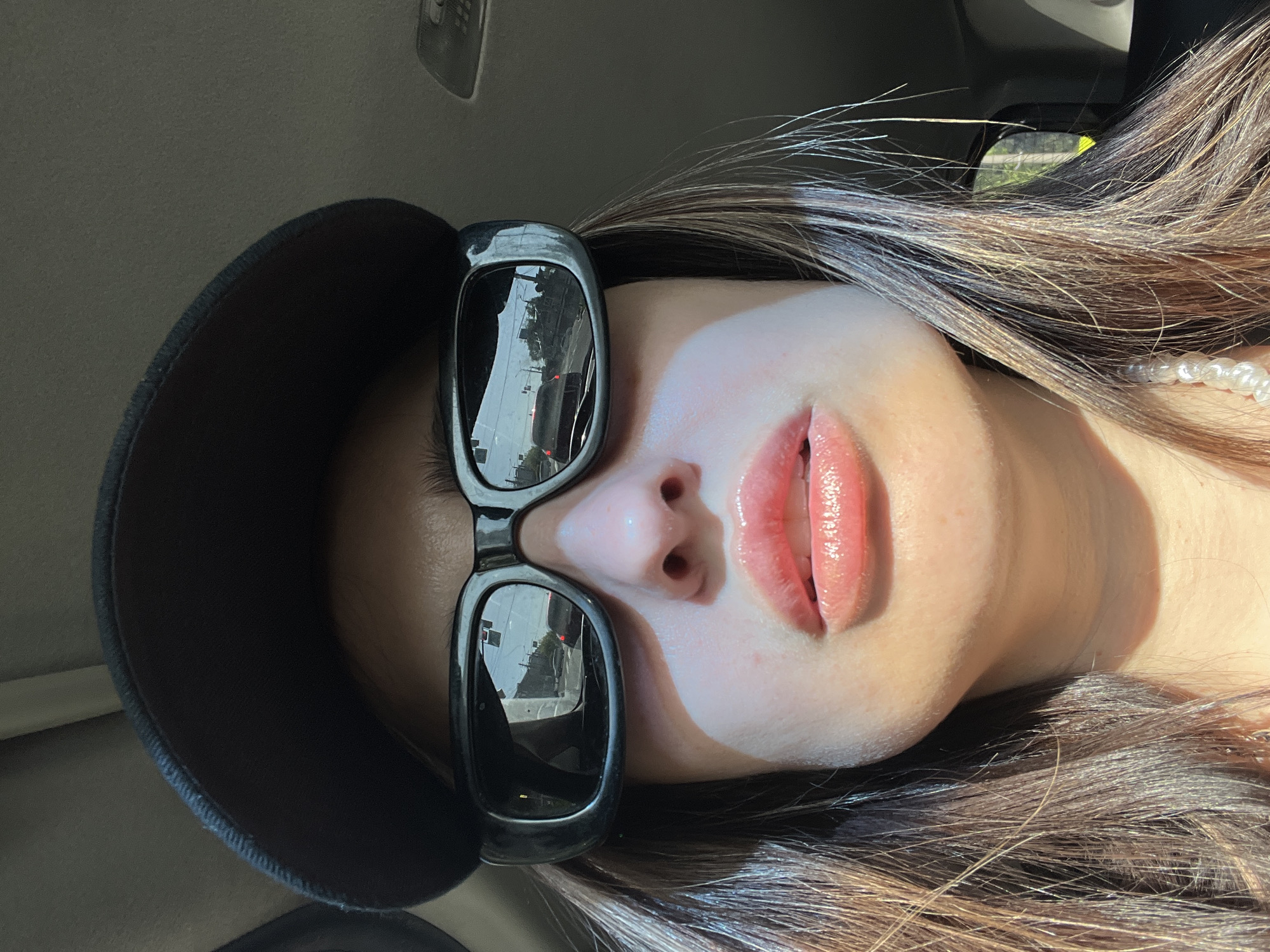 A medium shot depicts a person wearing sunglasses and a black cap. The reflection in the sunglasses shows buildings and a car in the city of Unknown, Unknown. The person is positioned inside a car, with the car's roof visible in the upper left corner. A string of pearls is visible near the person's neck. The lighting suggests daytime.