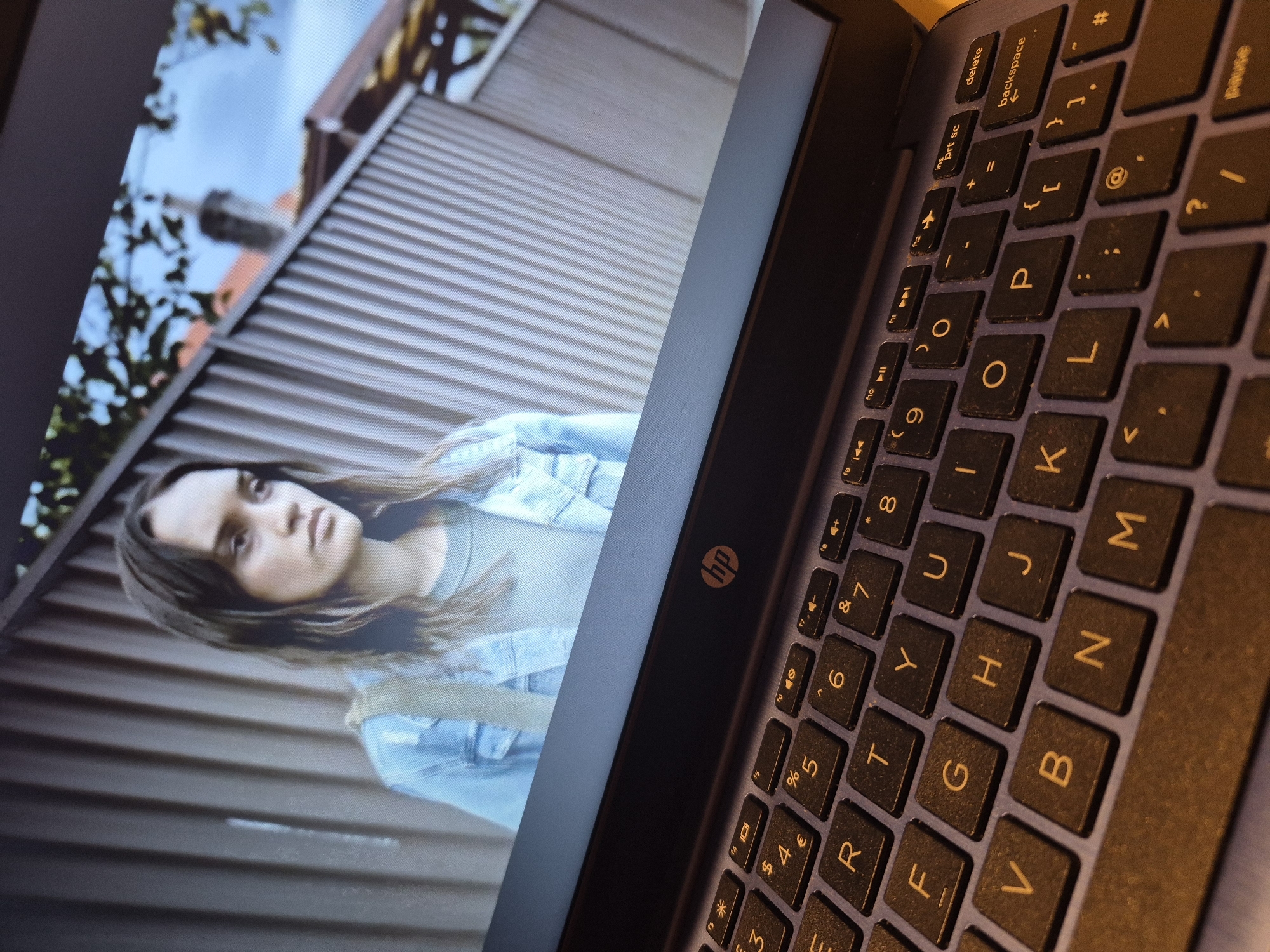 An HP laptop displays a young woman in a light blue denim jacket and grey t-shirt, looking to her right with a neutral expression. The background on the screen shows a corrugated metal fence and a hazy view of rooftops and trees under a blue sky. The laptop's keyboard is in focus in the foreground, showing a partial QWERTY layout with keys such as 'E', 'R', 'T', 'Y', 'U', 'I', 'O', 'P', 'A', 'S', 'D', 'F', 'G', 'H', 'J', 'K', 'L', and 'M', as well as number and symbol keys. The HP logo is visible on the laptop's bezel. The scene does not provide any specific information that can be used to determine a location in the city of Sandwell, United Kingdom.