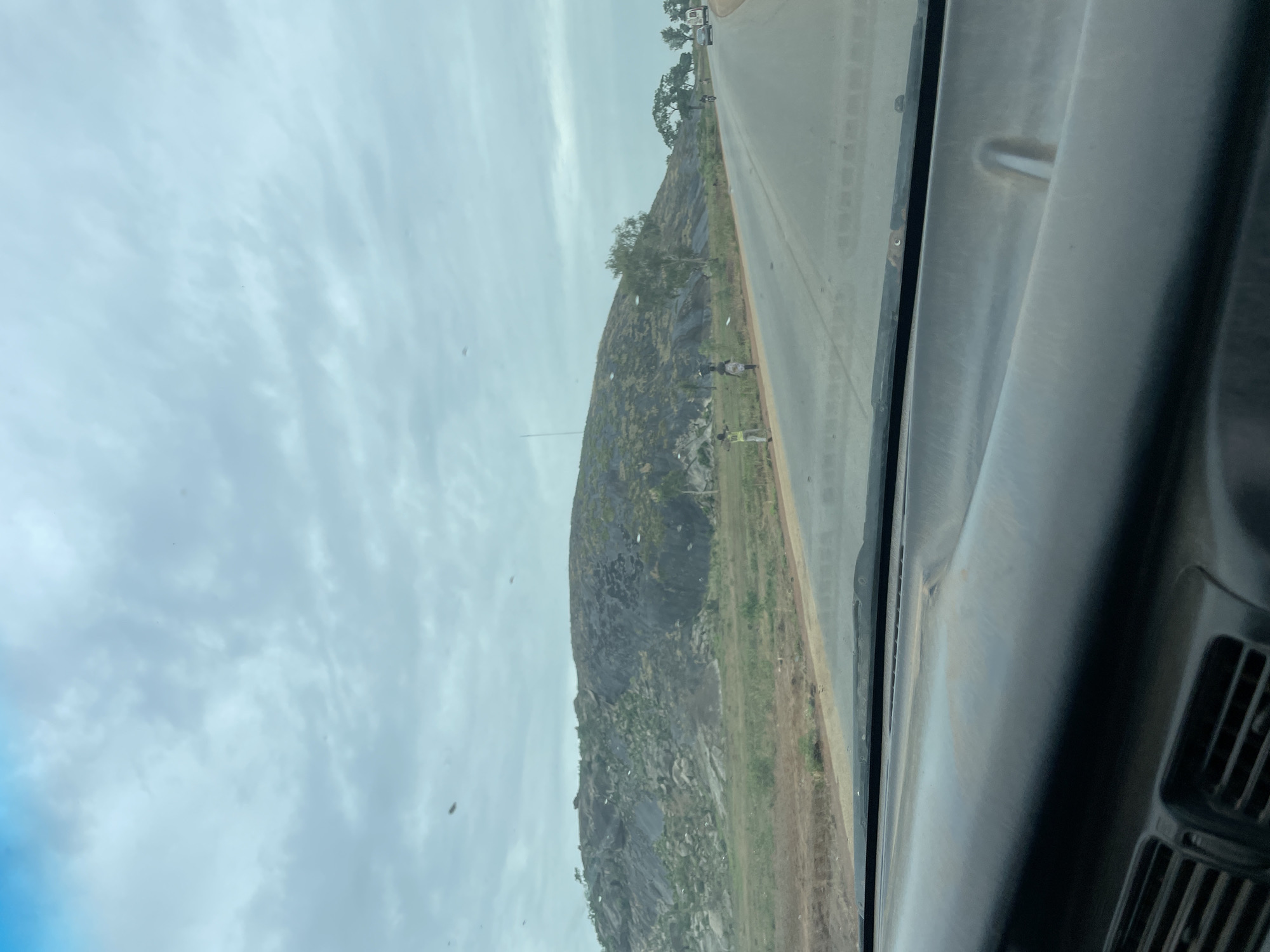A rocky outcrop rises beside a highway near Kujama, Nigeria. Two people stand by the roadside, one wearing a yellow vest. A few trees grow on the hill. A vehicle is driving on the highway. The sky is overcast. The photo was taken from inside a vehicle, looking through the windshield.