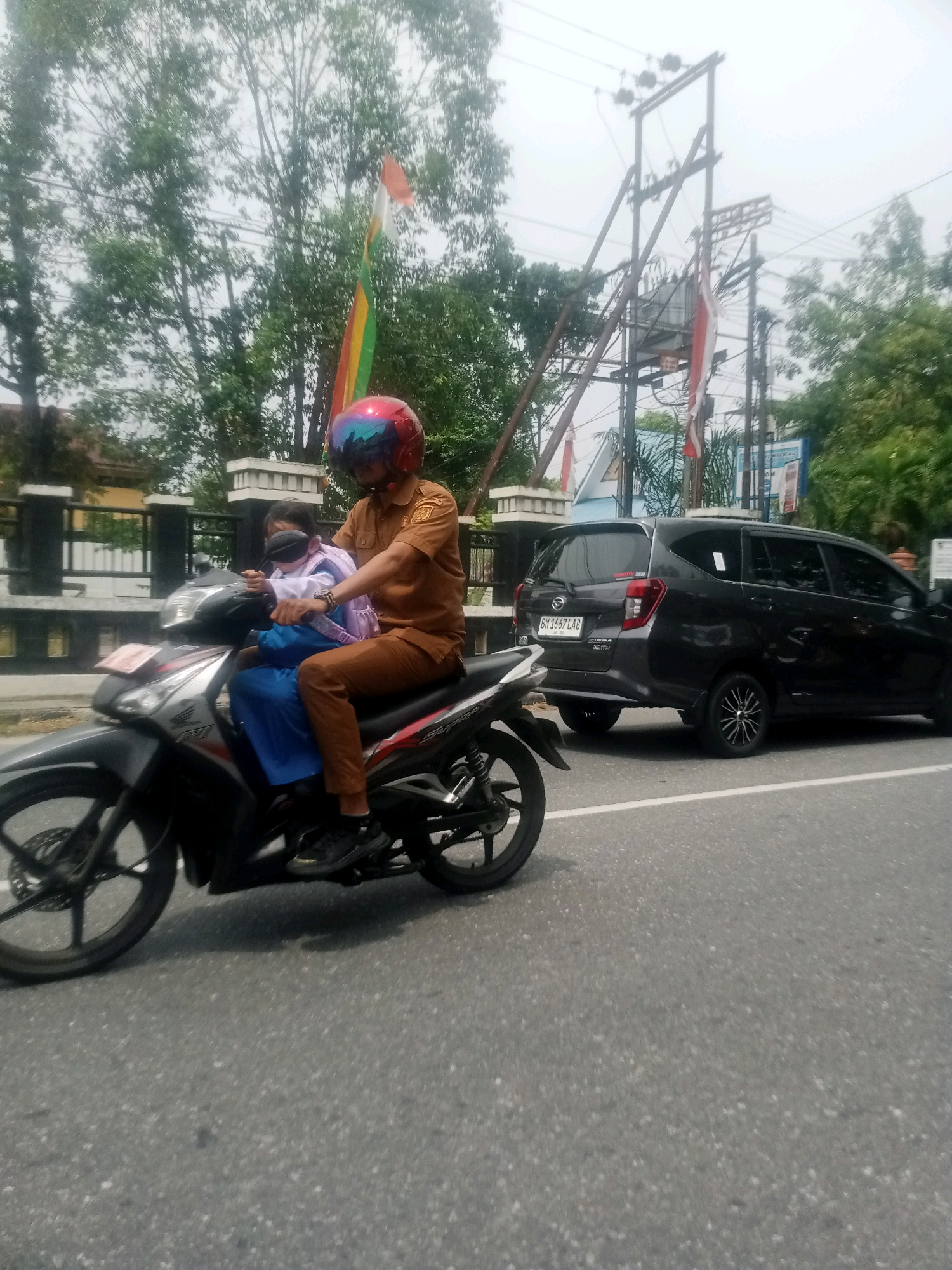 A man wearing a brown uniform and a red helmet rides a motorcycle with a child in front of him. The child is wearing a white shirt, a blue skirt, and a backpack. The man is driving on a road in Pekanbaru, Indonesia. In the background, there are trees, power lines, and a black car. The sky is cloudy.