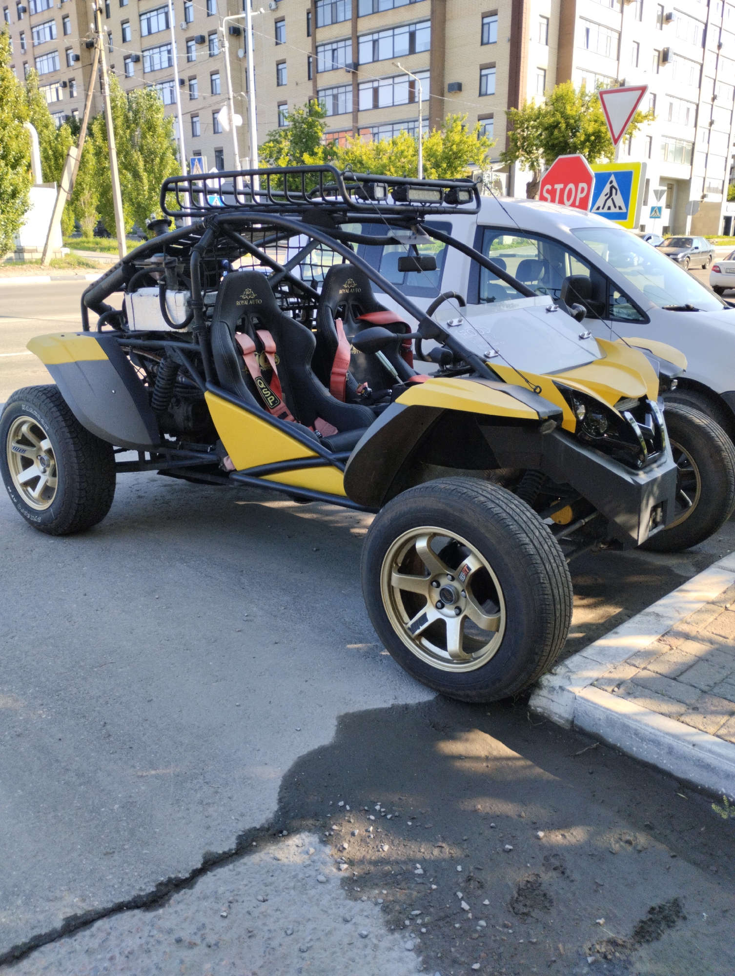 A yellow and silver off-road buggy is parked on an asphalt road next to a sidewalk with a concrete curb. The buggy has a roll cage, racing seats with red harnesses, and gold alloy wheels. In the background, there are several apartment buildings, trees, power lines, and traffic signs, including a stop sign and a pedestrian crossing sign. The scene appears to be a street in the city of Kostanay, Kazakhstan.