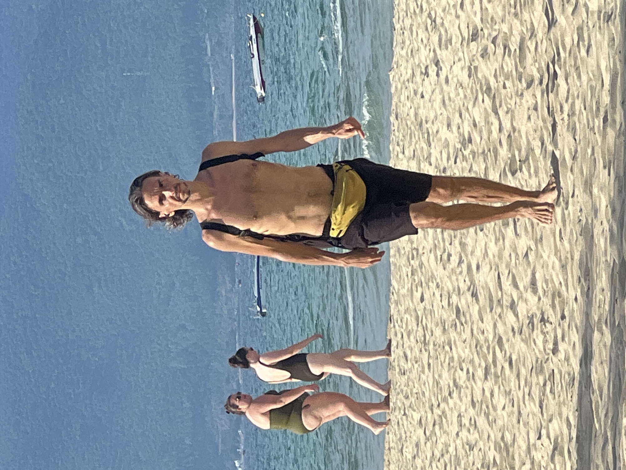 A man with long, dark hair and a lean physique walks barefoot on a sandy beach in Đà Nẵng, Vietnam. He is shirtless, wearing black shorts and a yellow fanny pack around his waist. Black straps from a backpack are visible over his shoulders. Two women in swimwear are walking away from him on the beach to his left. The woman closer to the foreground wears a dark swimsuit, and the woman further back wears a dark olive swimsuit. The ocean is visible behind them, with a few boats on the water and a hazy, mountainous landscape in the distance. The sky is clear and blue.