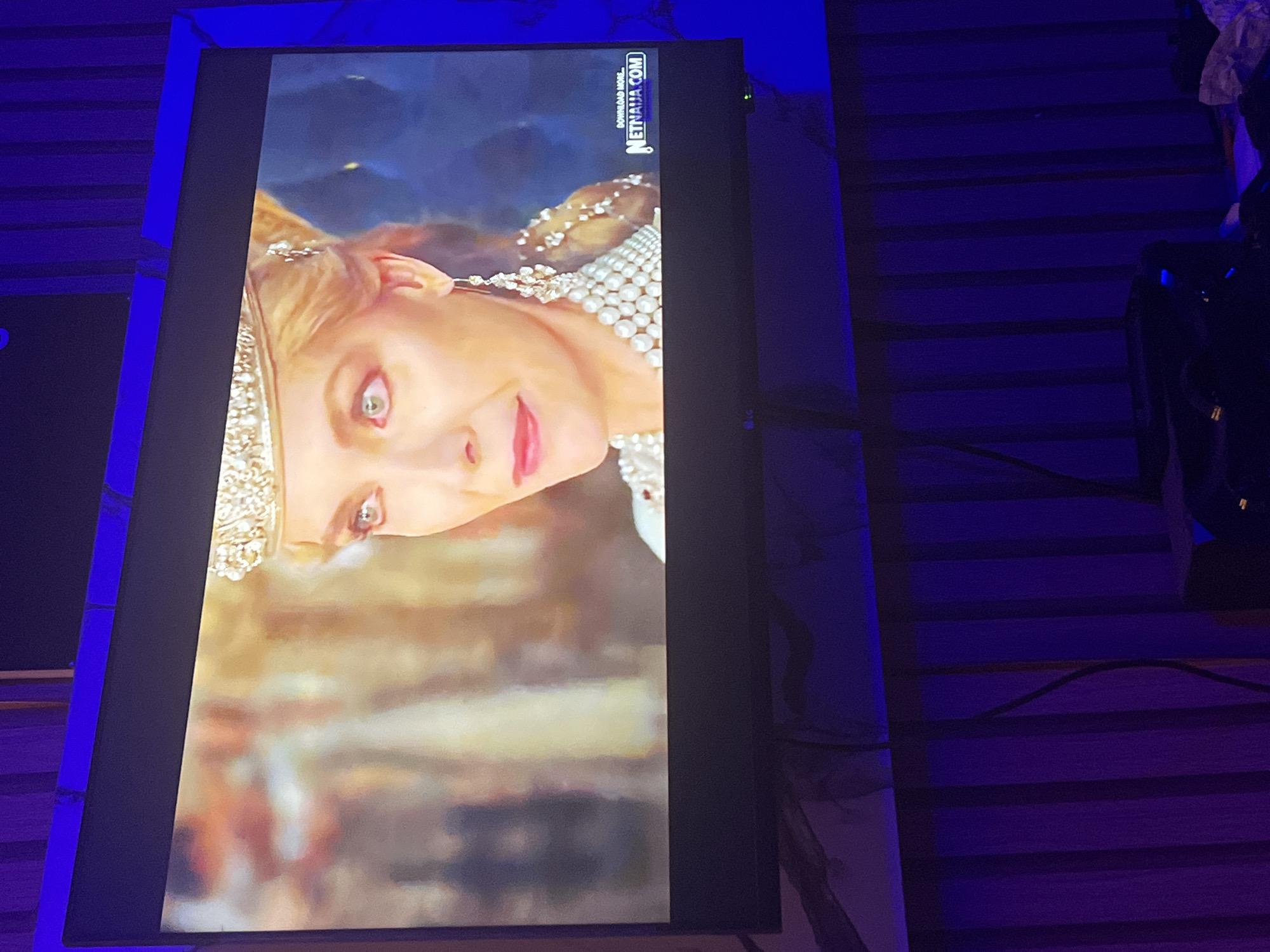 A rectangular screen, positioned vertically and tilted slightly, displays a still frame of a fair-skinned woman. The woman has light-colored hair, green eyes, and pink lips, wearing an elaborate golden crown or headpiece and a pearl-encrusted garment or necklace. She sports a subtle, enigmatic smile. The background on the screen appears to be a blurred landscape or abstract imagery.
The room surrounding the screen is dark, illuminated predominantly by a strong blue-purple light. Horizontal lines, possibly from a paneled wall or blinds, are visible on the wall behind the screen. To the right, some dark, indistinct objects are present.
Visible text on the top right portion of the screen (when the screen content is viewed upright) reads "DOWNLOAD MORE" and "NETNAIJA.COM", oriented vertically. The image captures a moment of media consumption in a dimly lit indoor setting.