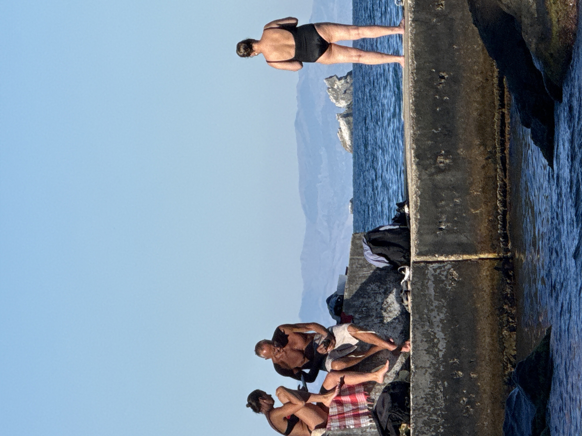 In Partenit, Ukraine, three individuals are present on a concrete pier under a clear blue sky. On the left, a woman in a bikini and a man wearing shorts are seated, with the man holding a dark object. Nearby are a red and white checkered towel and black bags. To the right, a woman in a black one-piece swimsuit stands facing away from the camera, looking towards the sea. In the distance, rocky outcrops emerge from the blue water, with hazy mountains forming the backdrop. The sea is calm, and the pier is constructed of weathered concrete.