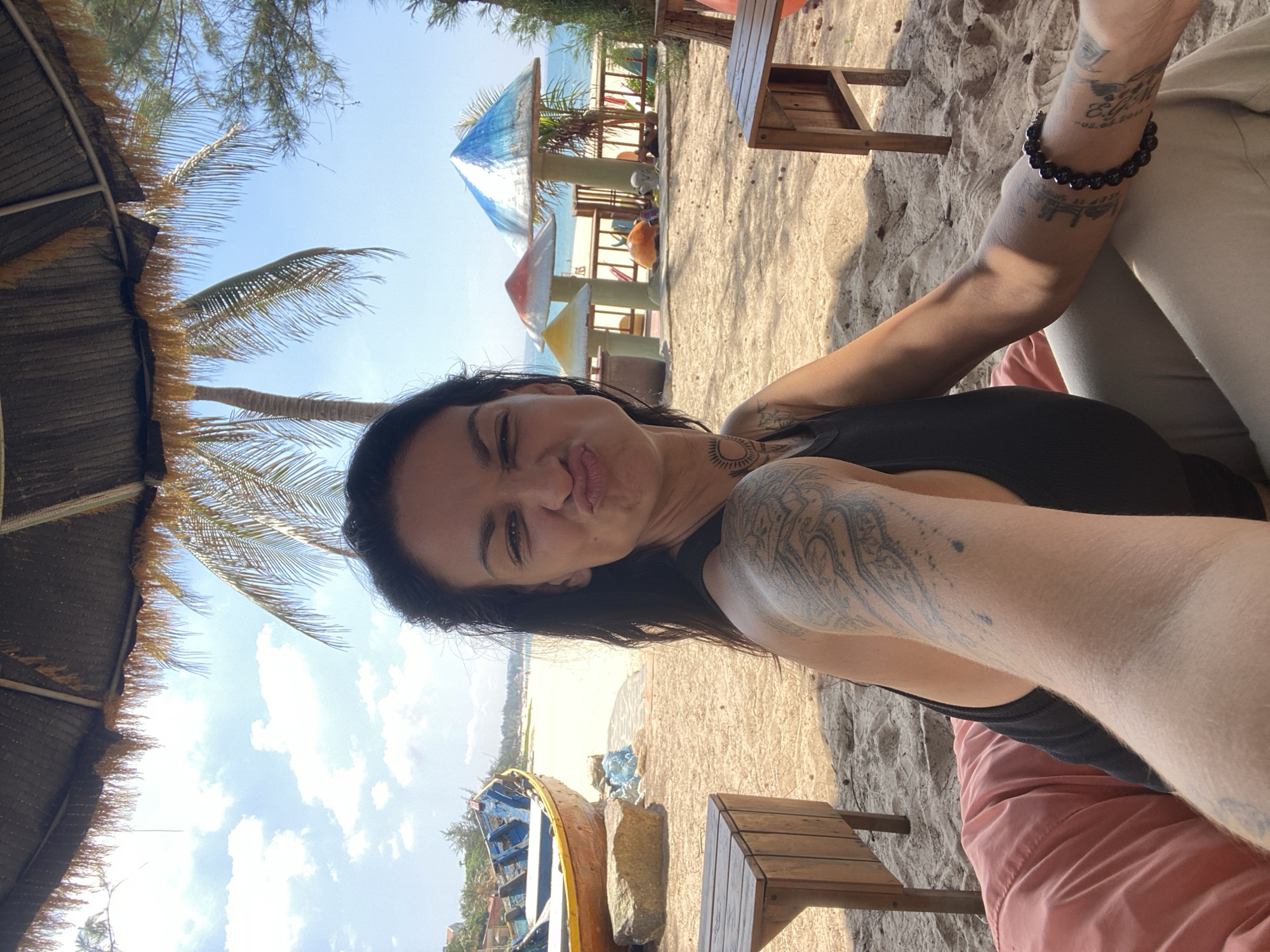 A woman is taking a selfie on a sandy beach in Phu Thuy Ward, Vietnam. She is wearing a black tank top and light-colored pants. She has numerous tattoos on her arms and upper back. The woman is pouting her lips and looking at the camera. In the background, there is a thatched umbrella, palm trees, and a blue and yellow boat on the sand. There are also colorful mushroom-shaped structures and the ocean visible. The scene appears to be a sunny day, with a clear blue sky and scattered clouds.