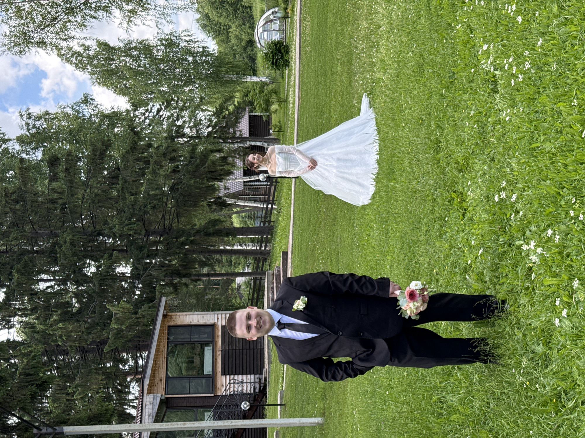A man in a black suit and tie stands in the foreground, holding a bouquet of pink and white flowers, with his left hand in his pocket. He is smiling at the camera. In the background, a woman in a white wedding dress stands with her hands clasped in front of her, looking to her left. They are in a grassy field with scattered white flowers. Behind them are trees, a wooden building with a peaked roof, and a geodesic dome structure. The image appears to be taken in Votkinsk, Russia, given the context provided. The scene suggests a wedding or pre-wedding photoshoot.