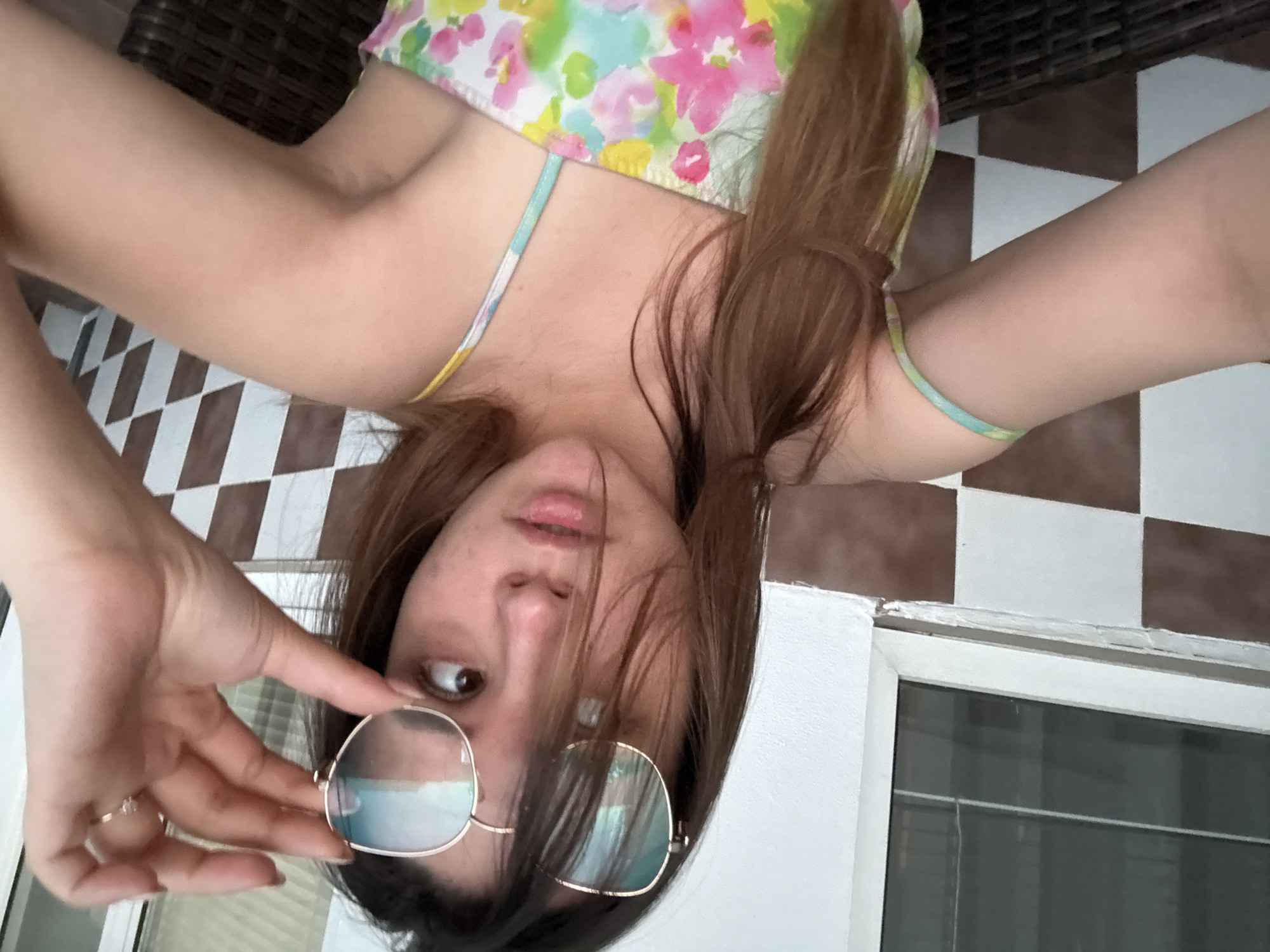 A young woman is depicted in a self-portrait or selfie. She is seen from an upside-down perspective. The woman is holding a pair of round-framed sunglasses in her left hand, partially obscuring her face. She has brown hair and is wearing a floral-patterned top with a thin strap. Her skin tone appears fair. A ring is visible on one of her fingers. The background includes checkered tiling and a white wall, suggesting an indoor setting possibly in a building. The location of Sofia, Bulgaria, is not identifiable based on the provided information alone.