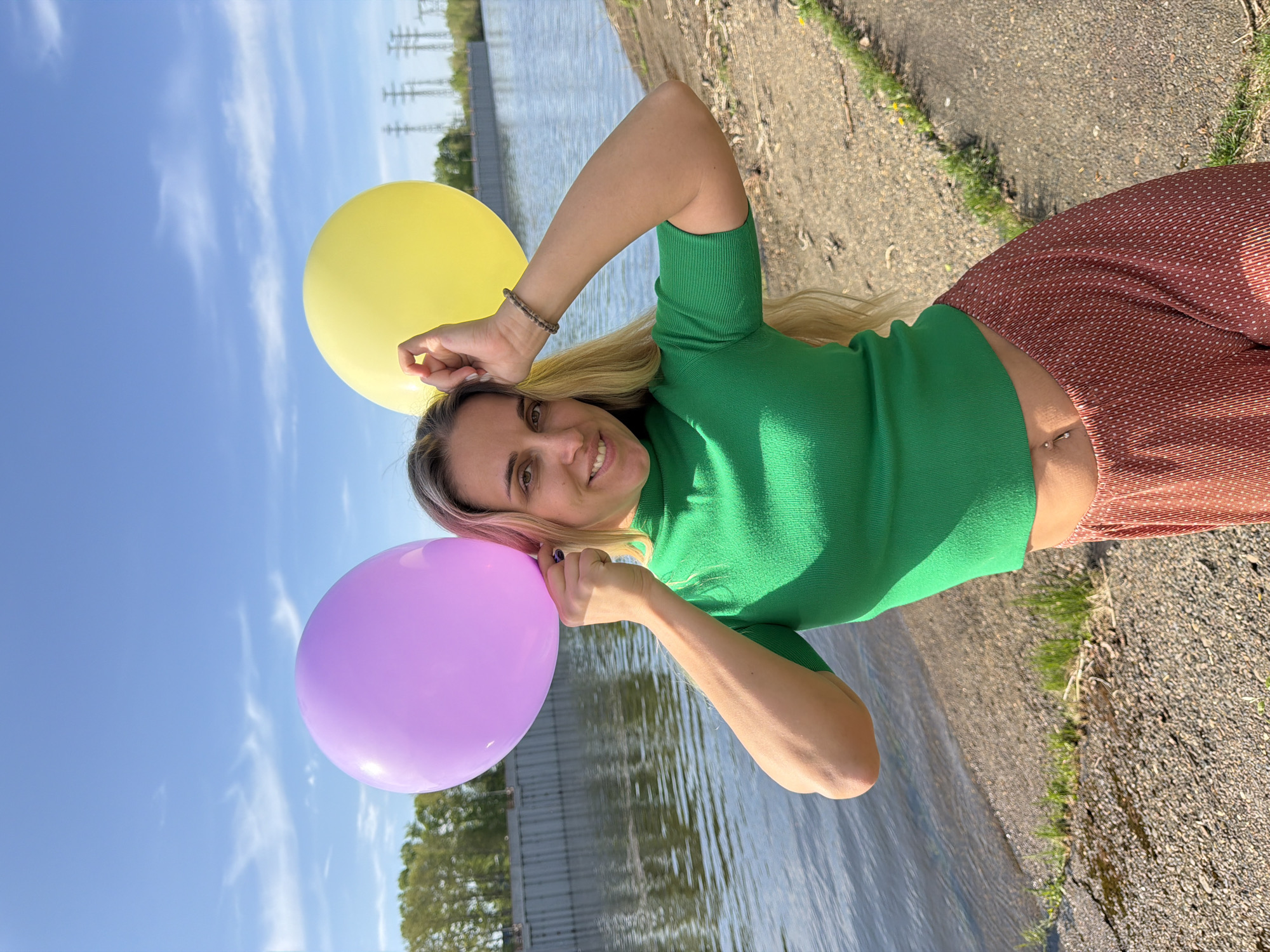 A fair-skinned woman with long blonde hair stands outdoors on a stone and grass embankment next to a body of water. She wears a green, short-sleeved shirt and patterned brown pants. In her hands, she holds a yellow and a purple balloon. The location appears to be Chaikovsky, Russia, as evidenced by the power lines visible in the background across the body of water.