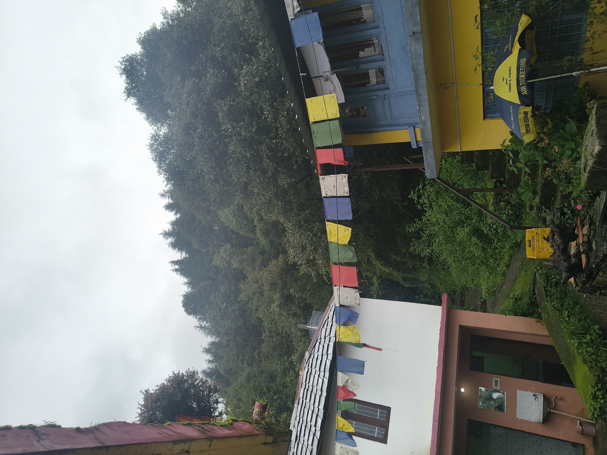 The image depicts a cloudy day in a mountainous region. In the foreground, two buildings are visible. The building on the left is white with a pink trim and a grey tiled roof. Prayer flags in various colors (red, blue, yellow, white, and green) are strung across the front of this building. A window with a dark brown frame is visible on its upper story. Below, an open-air area features a sink, a mirror, and two doors, one of which is partially visible.
The building on the right is painted yellow on its lower story and light blue on its upper story, with blue window frames. Prayer flags are also strung across the front of this building. A yellow and blue umbrella with the text "TOURS & TRAVEL", "AIR TICKETING", and "FOREIGN EXCHANGE" is open and supported by a white pole.
Behind the buildings, a lush green hillside covered in dense trees rises steeply. The sky is overcast, suggesting a possibly humid or rainy atmosphere. A stone staircase is visible ascending the hill between the two buildings. A yellow plastic crate is placed on the ground to the right of the staircase. The overall impression is of a rural or semi-urban setting nestled within a natural, forested landscape. The location is identified as Haini, India.