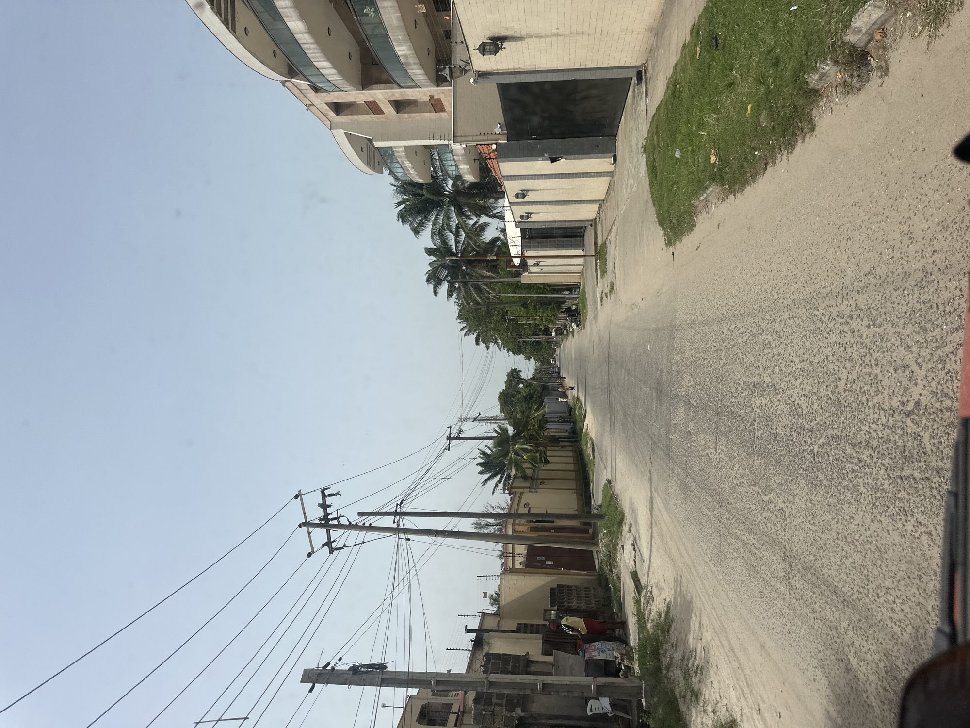 The image depicts a street scene in Lagos, Nigeria, characterized by a narrow road lined with buildings of varying sizes and architectural styles. The road surface appears to be concrete or asphalt, showing signs of wear. Buildings range from modern multi-story structures to smaller, older constructions, indicative of diverse urban development. Palm trees and other greenery are interspersed along the street, adding a touch of tropical ambiance. Overhead power lines and utility poles create a visual network in the sky. In the foreground, a portion of a vehicle is visible, suggesting the image was taken from inside a moving car or similar transport. The overall atmosphere is indicative of a typical residential or commercial area in Lagos, with its blend of structures and natural elements.