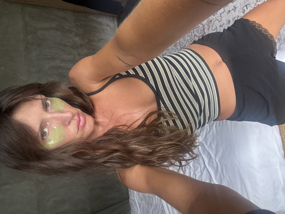 A woman lies on a white bed in a selfie. She wears a striped tank top and black shorts with lace trim. Two light green under-eye patches are on her face. Her brown hair is styled in waves. The setting appears casual, possibly a bedroom. Her right arm is extended towards the camera, while her left arm rests along the bed.
