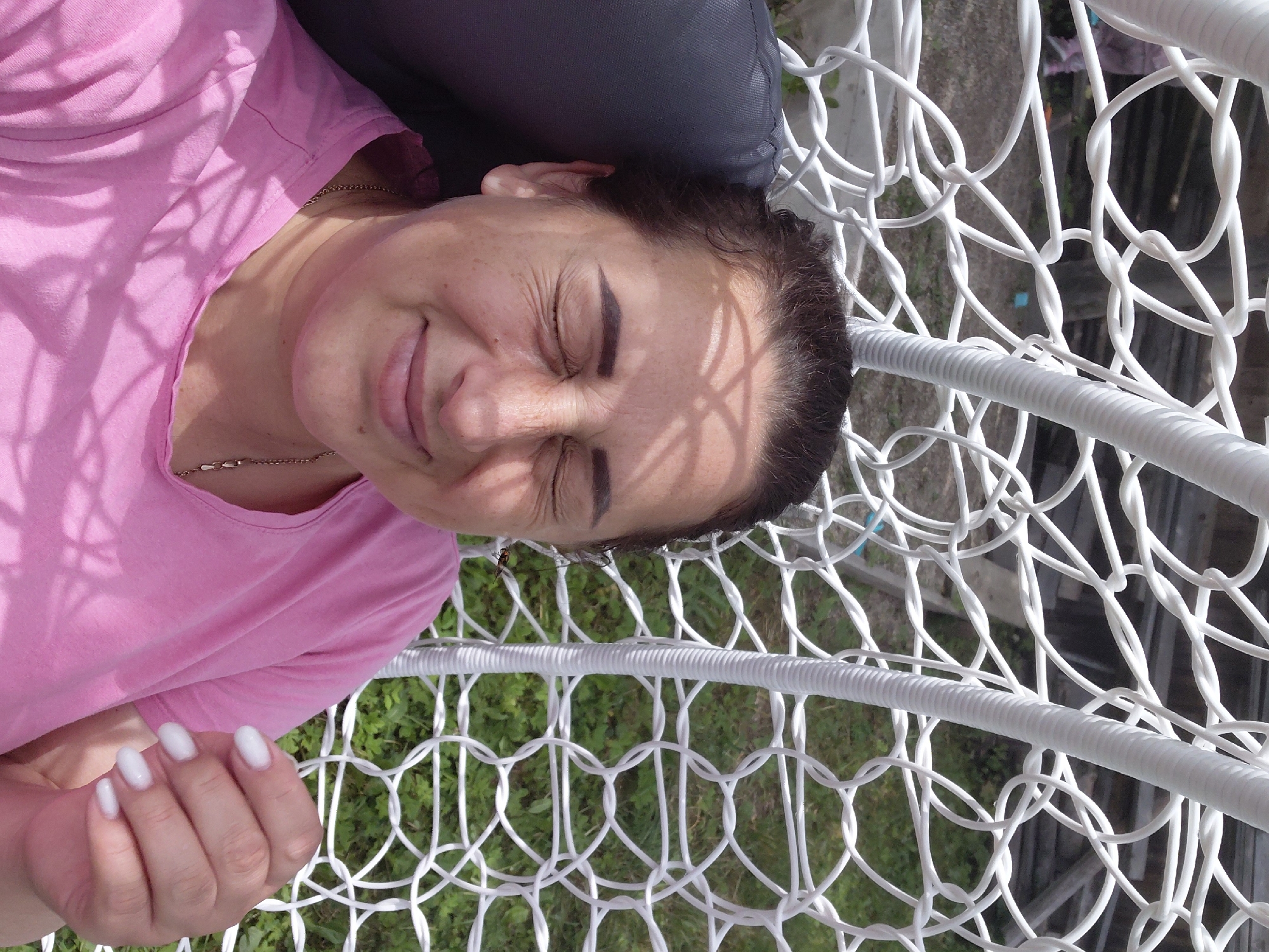 A woman with brown hair and closed eyes is lying in a white woven hanging chair. She is wearing a pink t-shirt and a gold necklace. Her fingernails are painted white. Shadows from leaves are cast on her face and shirt. The background shows green grass and a wooden structure. The image is taken outdoors on a sunny day.