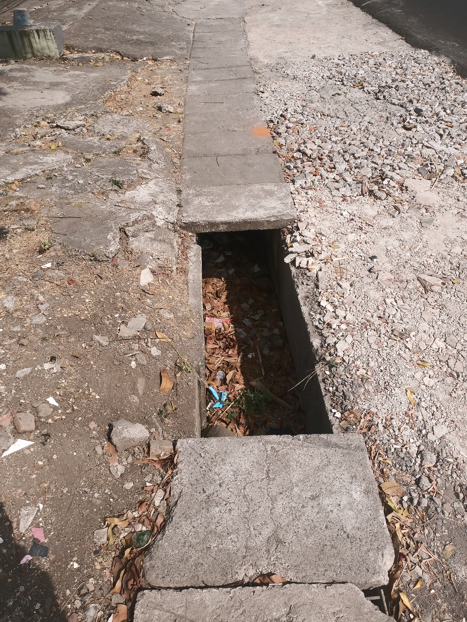 The image depicts a section of a concrete drainage ditch filled with debris, including fallen leaves, small rocks, and a few pieces of plastic. The ditch is located alongside a cracked concrete pathway and a gravel-covered area. The environment suggests an outdoor urban setting, possibly a street or sidewalk in the city of Malang, Indonesia, given the context of the request. The lighting indicates it is daytime.