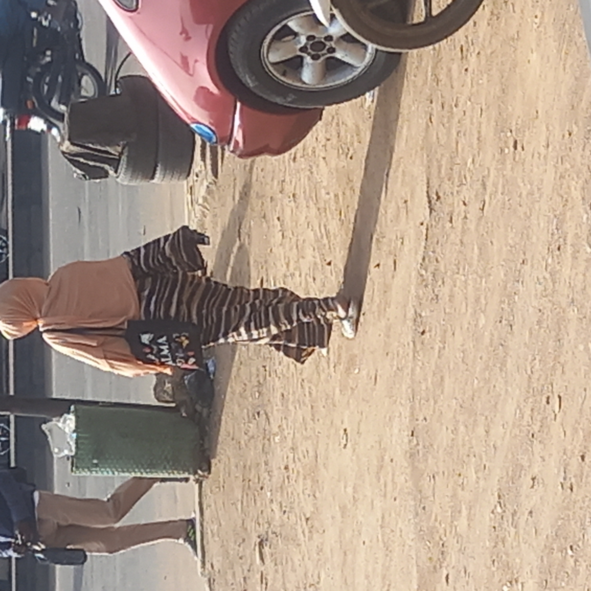 An outdoor scene captures a woman walking on a dusty, unpaved roadside, likely in Dadi Riba, Nigeria. The woman, positioned centrally in the frame, is seen from the side, wearing a light peach or tan head covering and a matching loose top. She is also dressed in vertically striped black and brown/beige trousers or a long skirt. Over her shoulder, she carries a black tote bag with the word "ALMA" clearly visible in white text, accompanied by colorful, abstract floral-like designs. She wears white shoes, and a prominent long shadow is cast from her figure onto the ground, indicating bright sunlight, characteristic of either early morning or late afternoon.
On the far left, the lower body of another person is partially visible, wearing brown trousers and dark sneakers with white soles, suggesting they are also walking or standing nearby. In the background, a red car is partially visible, showcasing its front wheel with silver spokes and a portion of its body. Behind the car, a dark-colored motorcycle or bicycle is indistinctly seen. A rectangular green container, possibly a trash bin, rests near the lower-left edge of the frame. The ground is a light brown, earthy, and granular texture, consistent with an unpaved road or pathway.