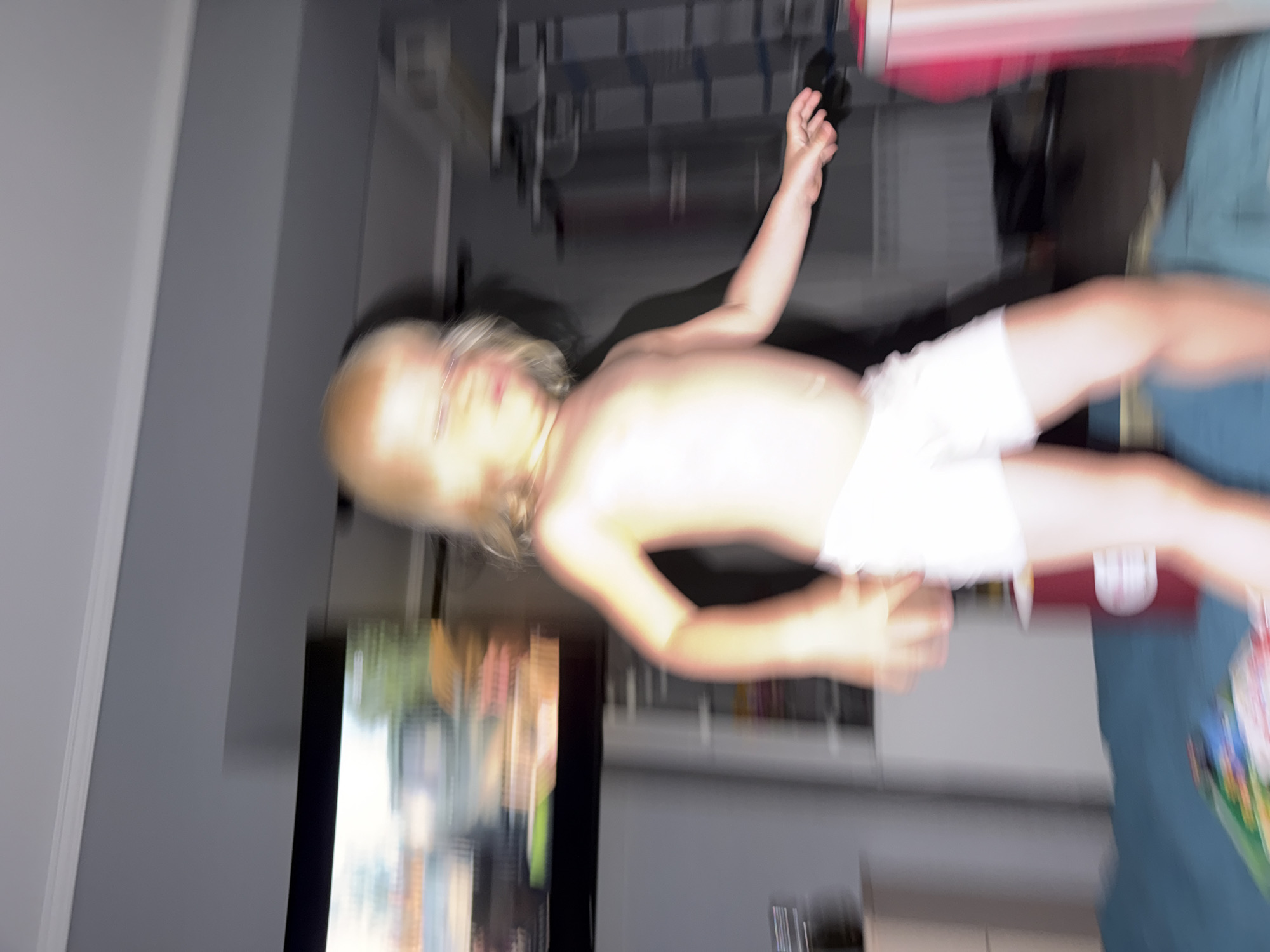 The image shows a blurred depiction of a young child, likely a toddler, standing on what appears to be a bed or soft surface. The child has blonde hair and is wearing white shorts. The child's body is partially obscured by motion blur. In the background, a television screen is visible, displaying a colorful image, and there are other indistinct objects in the room. The lighting appears to be artificial and warm. The room's walls are a neutral gray color. The overall impression is of a candid, possibly playful, moment captured indoors. The image does not contain any discernible features that would allow for identification of the specific location within Sochi, Russia.