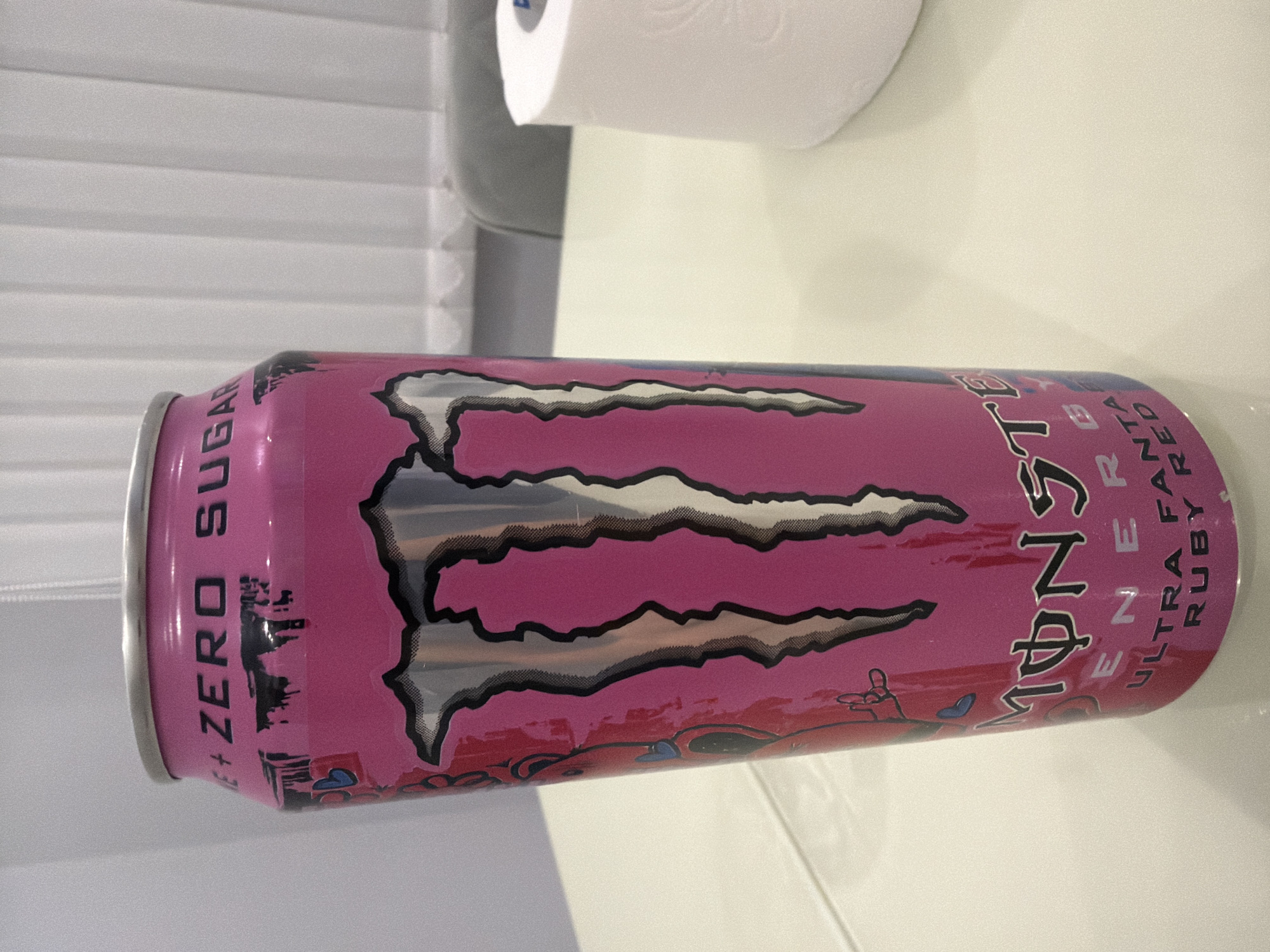 A pink Monster energy drink can, labeled "ZERO SUGAR ULTRA FANTA," sits on a white table. The can features the iconic Monster logo in silver and black. To the right of the can, a roll of white toilet paper is partially visible. The background consists of white blinds and a gray chair, suggesting an indoor setting. The scene appears to be a casual domestic environment, likely in a home. The location is not specified to be in Novi Iskar, Bulgaria.