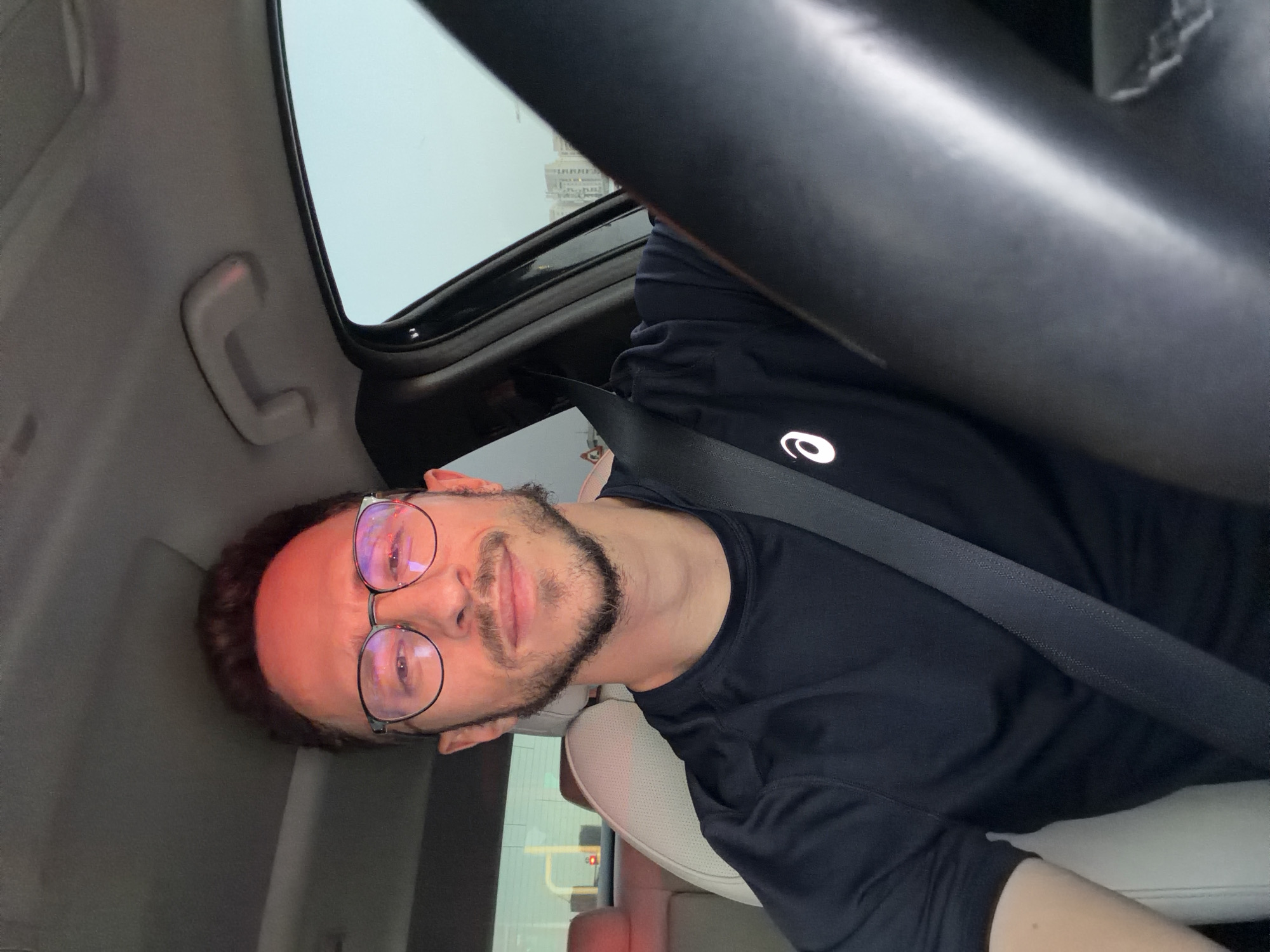 A man is in the driver's seat of a car. He has brown hair, a beard, and is wearing glasses and a black T-shirt with a logo. He is wearing a seatbelt. A building is visible through the car's sunroof. Based on the style of architecture, the image was likely taken in Tel-Aviv, Israel.