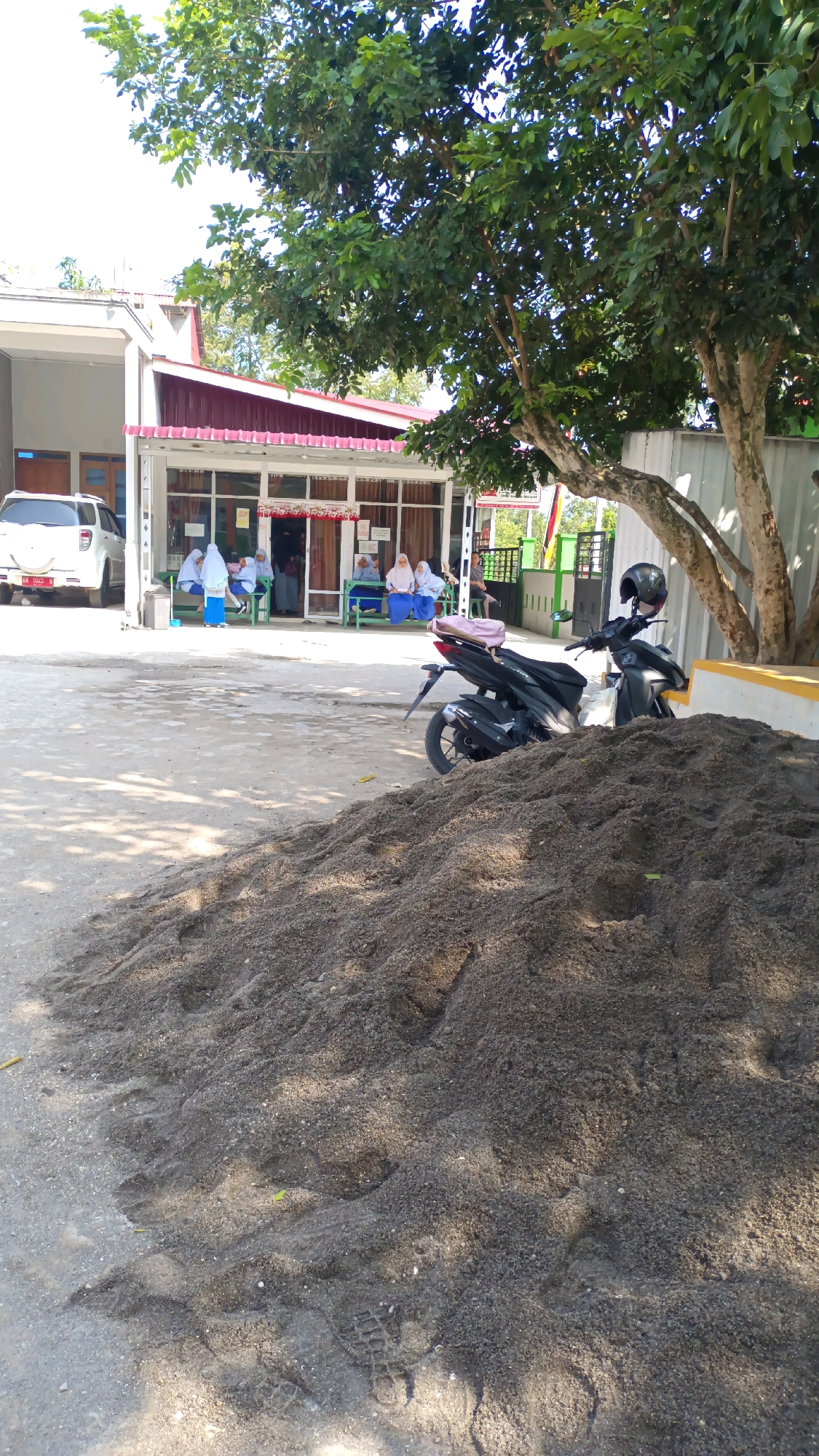 In Padang Panjang, Indonesia, a scene unfolds outside a building with a prominent maroon awning and large glass windows. Several individuals, mostly women wearing hijabs, are seated on green benches. To the left, a white SUV is parked. In the foreground, a large pile of dark sand dominates the view. A black motorcycle with a helmet on its handlebars is parked on the pavement near the sand pile. The environment appears to be outdoors, possibly a public space or the grounds of an institution, under the dappled shade of a large tree.