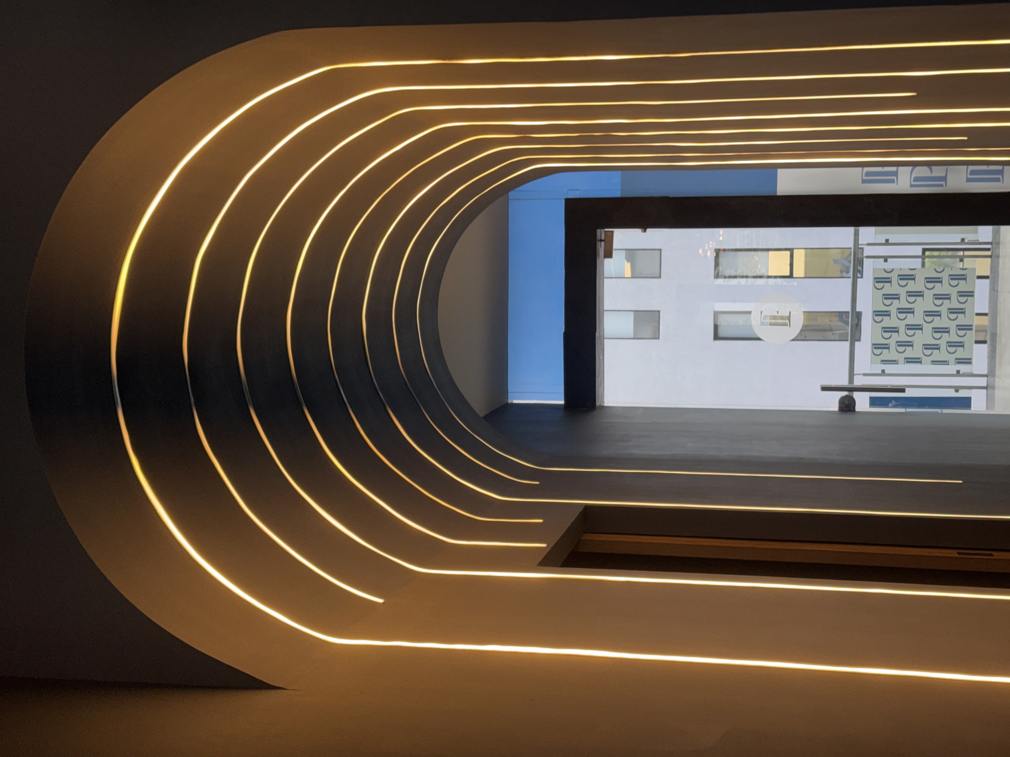 The image displays a modern architectural interior in Puebla, Mexico, featuring a dramatic, curved tunnel-like structure with integrated lighting. The dominant subject is the interior's left wall and ceiling, which are dark gray and adorned with numerous parallel, linear LED lights emitting a warm, golden-yellow glow. These lights follow the sweeping curve of the structure, creating a sense of depth and movement. The right side of the interior transitions into a flat, dark gray floor, also with embedded linear lights extending forward.
This illuminated passage opens up to a large window or doorway, revealing an exterior scene. Beyond, a contemporary white building with multiple rectangular windows is visible under a clear blue sky. On one of the exterior windows of this building, a circular logo is present, containing a stylized lowercase "e" in its center and the word "iNTERMOD" around its perimeter. Adjacent to this, a window film or banner features a repeating pattern of stylized lowercase "b" characters.
The setting appears to be a commercial or public space, designed with a focus on modern aesthetics and dramatic lighting. The clear blue sky indicates it is daytime. No people are visible, leaving the architecture and its striking illumination as the central focus.