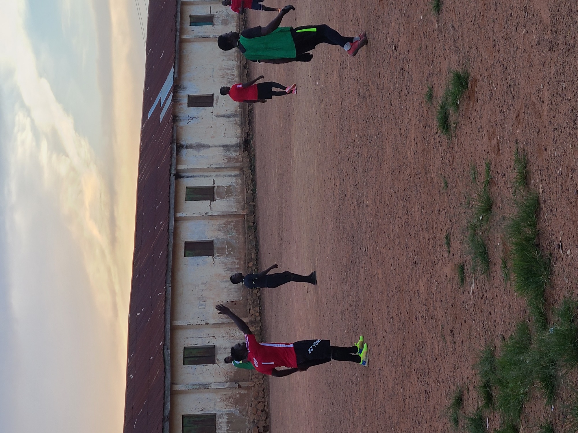 The image depicts a group of young men playing soccer on a dirt field in Jos, Nigeria. The scene is set against the backdrop of a long, weathered building with a rusty corrugated iron roof and several boarded-up windows. The sky above is a pale orange and blue, suggesting either sunrise or sunset. The field is predominantly brown dirt with patches of sparse green grass.
In the foreground, a man in a red jersey with the number '1' and black shorts is gesturing with his raised right arm. He is wearing bright yellow and green soccer cleats and dark socks. To his left, another man in a green vest and black shorts is seen from behind, also in motion. Further back, several other men in red jerseys and black shorts are visible, engaged in the game. One man in a black jersey and shorts stands to the left, seemingly observing. The overall impression is of an informal soccer match taking place in an outdoor, possibly rural or semi-urban, setting within Jos.