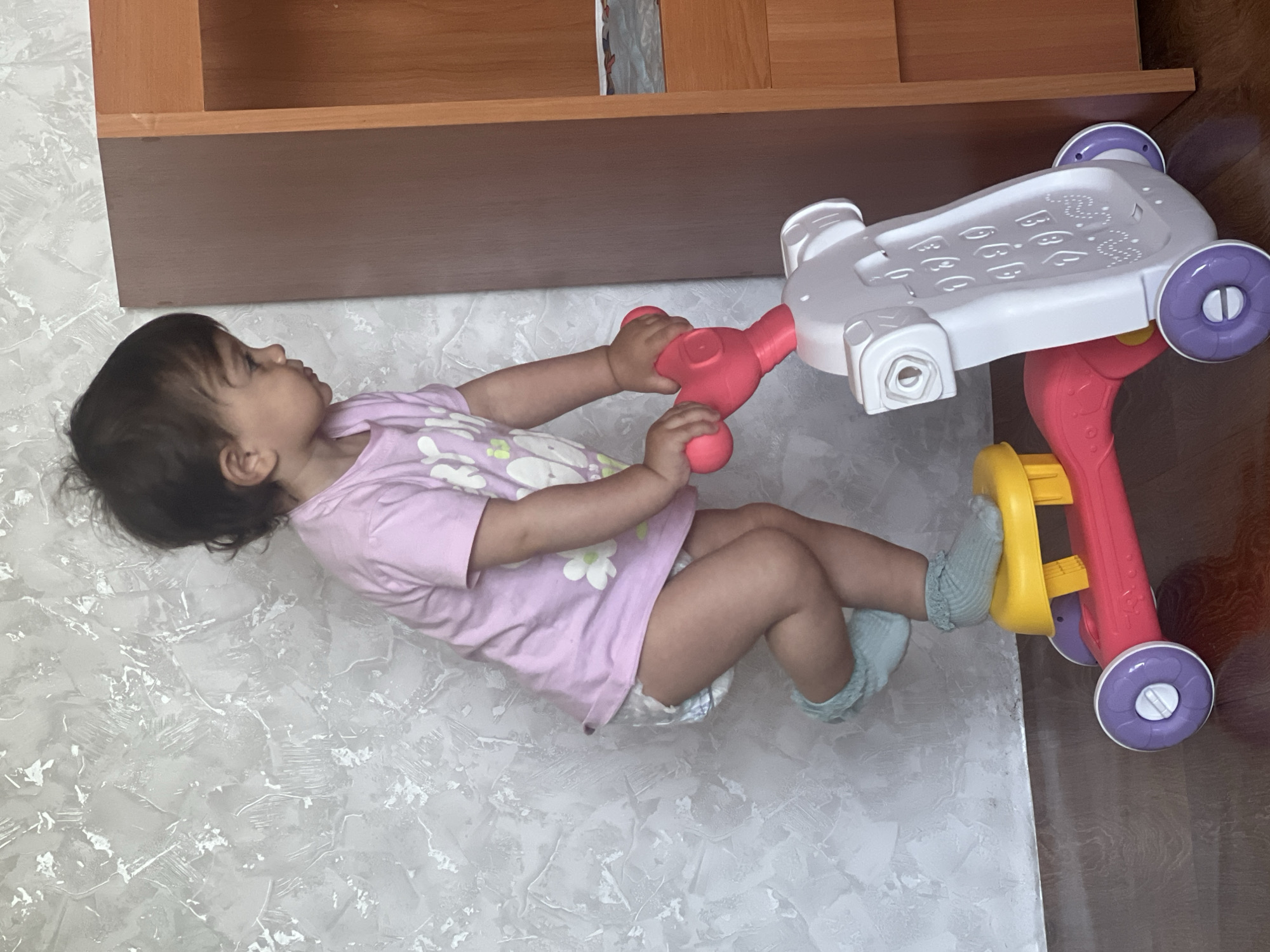 A toddler, wearing a pink t-shirt, a diaper, and light blue ruffled socks, stands on a toy scooter. The scooter has a pink frame, a yellow seat, and a white body with raised buttons resembling a keypad. The child is gripping the red handlebars. The scene takes place indoors, in what appears to be a room with a textured gray wall and a wooden floor. A portion of a brown wooden cabinet is visible on the right side of the image. The location is not specified in the image, and therefore it is not possible to determine if it is in Elena, Bulgaria.