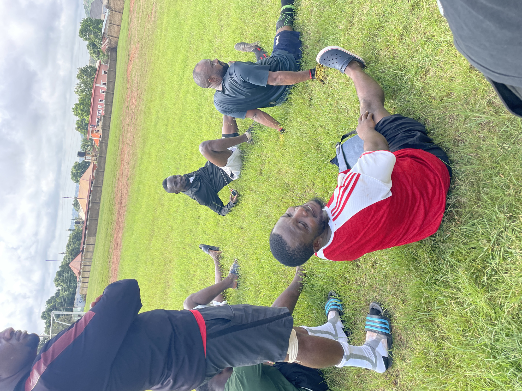 This image depicts a group of men in an outdoor grassy field, likely a sports ground, in Jos, Nigeria. The overcast sky suggests a cloudy day. In the foreground, a man in a red and white shirt and black shorts is seated on the grass, looking to his right with a smile. To his left, another man in black athletic wear is also seated, with his legs extended. Further back, two other men are seated on the grass, one in a black shirt and shorts, and another in a dark grey shirt and shorts, wearing athletic gloves. The background reveals a fence, some buildings, and scattered trees, characteristic of an urban or semi-urban environment. The overall scene suggests a relaxed moment, possibly during or after a sports activity.