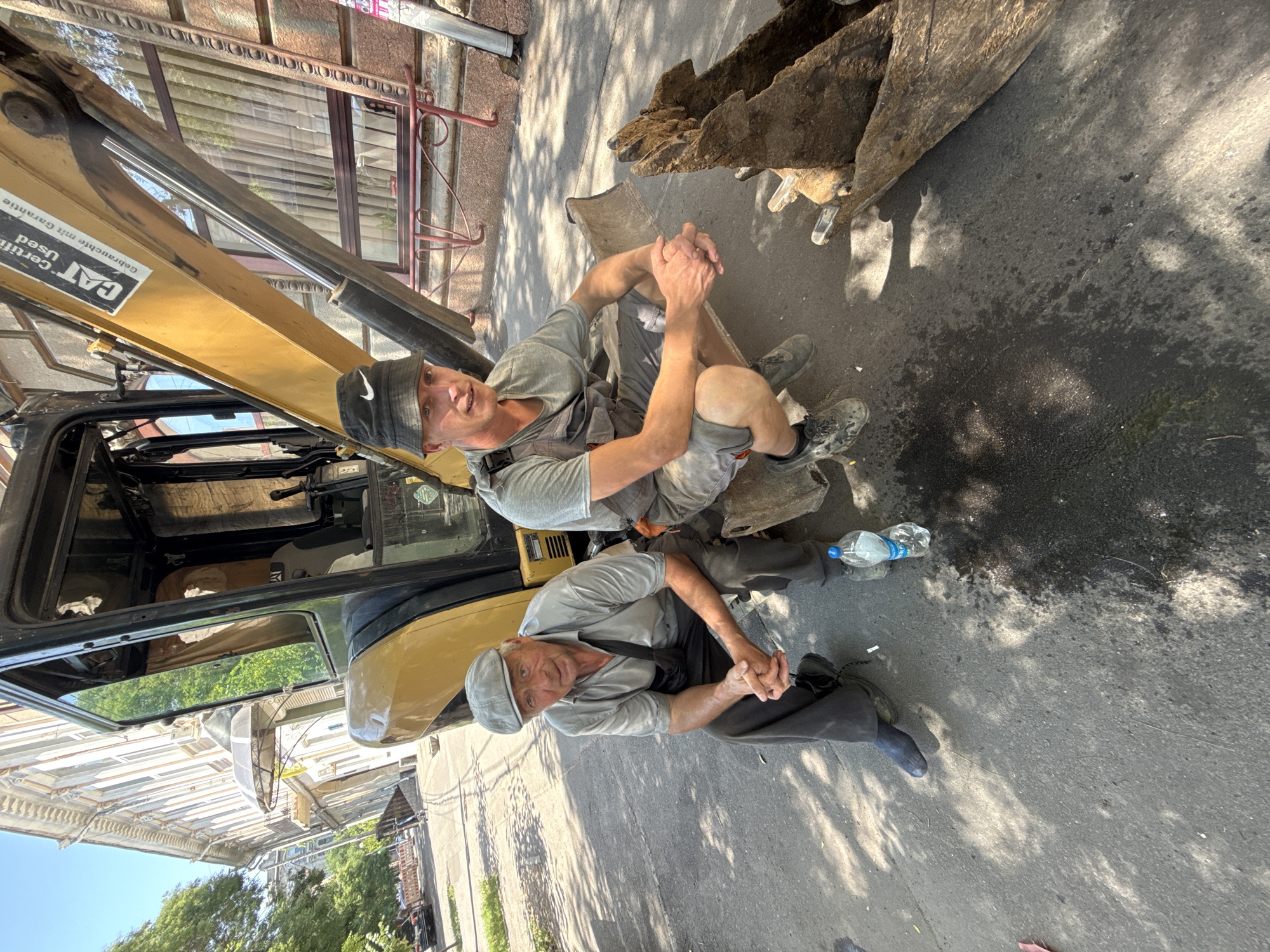 Two construction workers, one older with a flat cap and the other younger with a bucket hat, are seated on the bucket of a yellow excavator parked on an asphalt surface in an urban setting. The older man, wearing a light-colored short-sleeved shirt and dark pants, is looking towards the camera with his hands clasped. The younger man, in a gray t-shirt and overalls, is also looking at the camera with his hands clasped. A bottle of water is on the ground near them. In the background, a building with architectural details and trees line the street. The scene appears to be a construction site on a sunny day.