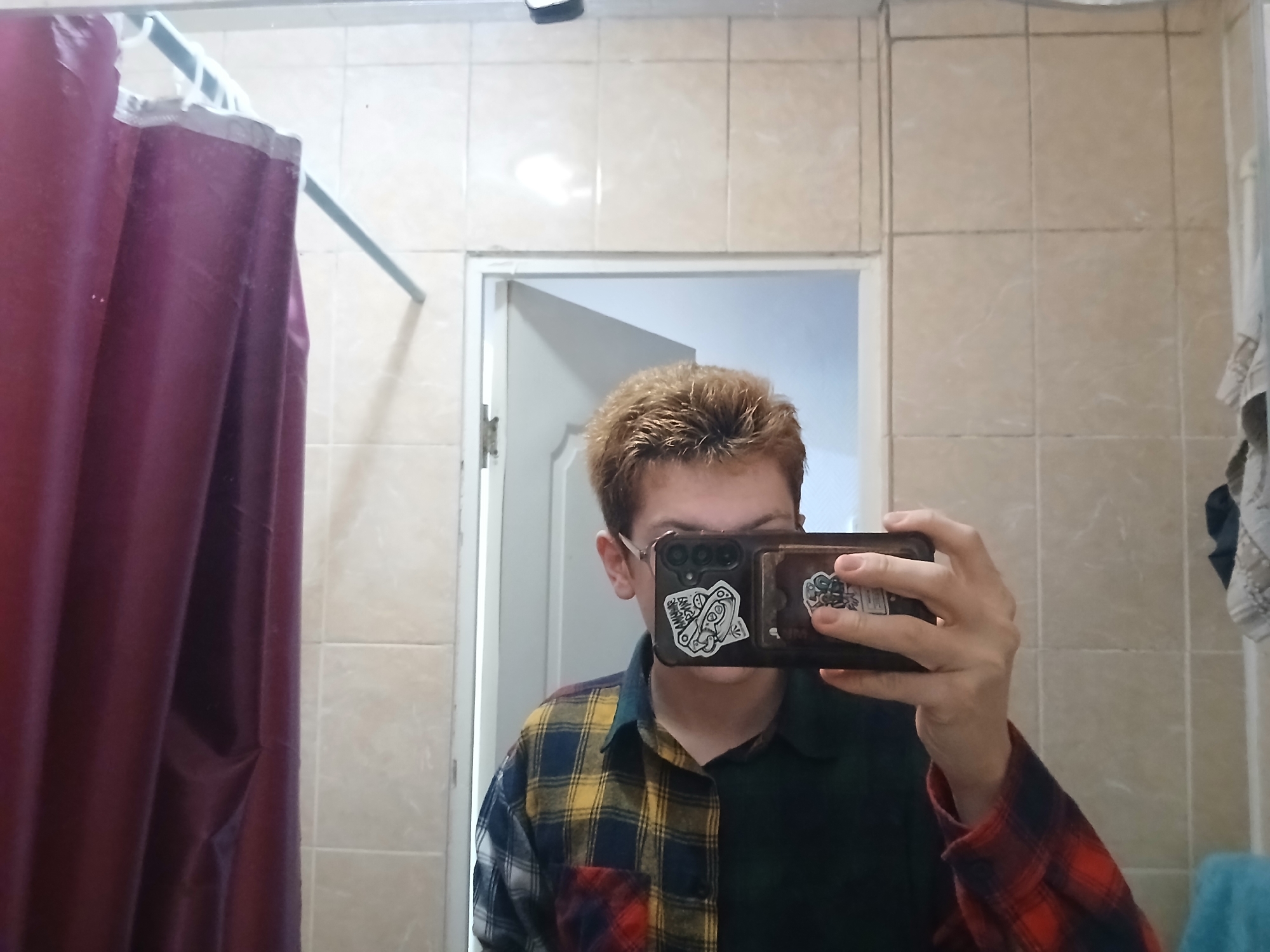A person is taking a selfie in a bathroom. The bathroom has light-colored tiles on the walls and a shower curtain on the left. The person is holding a smartphone with stickers on the case in front of their face. They are wearing glasses and a plaid shirt with mixed colors. The environment appears to be an ordinary bathroom setting. The location is in the city of Chaikovsky, Russia, but that information cannot be confirmed from the image alone.
