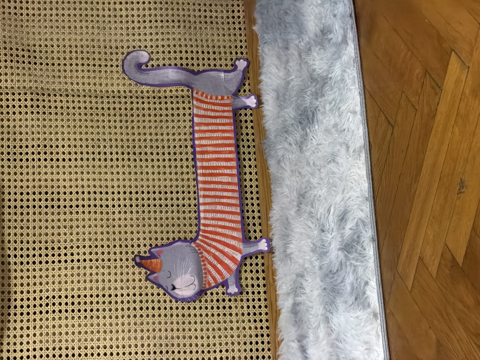 The image depicts a cartoon-like image of a gray cat wearing a striped orange and white sweater laying on what appears to be a woven wicker surface. The background includes a white shag rug and a wooden floor with a parquet pattern. There is no indication of a location in Moscow, Russia, or any people in the image.