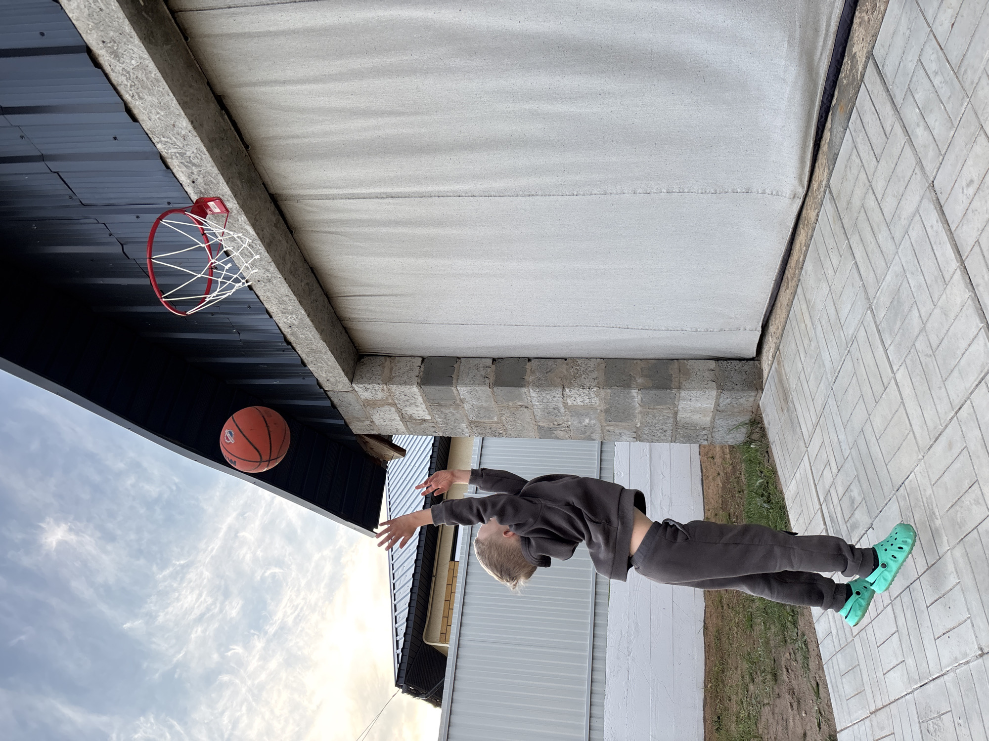This image captures a dynamic outdoor scene featuring a young boy engaged in playing basketball. The boy, who appears to be a child or pre-teen, is shown mid-leap, stretching upwards with both arms extended, seemingly attempting to score or reach for the ball. He is dressed in a dark grey tracksuit and distinctive bright turquoise Crocs. His blond hair is short and neat.
An orange basketball, with faint branding possibly reading "NCAA," is visible in mid-air, positioned slightly above and to the left of a red basketball hoop. The hoop, equipped with a white net, is mounted onto a dark, corrugated metal structure, which could be part of a shed or an outbuilding.
The ground where the boy stands is paved with light-colored, rectangular bricks arranged in a herringbone pattern. A narrow strip of bare earth is visible to the boy's right. Overhead, a large, light-colored fabric or tarp-like material acts as a ceiling or awning, supported by concrete beams and blockwork. In the background, beyond the dark metal structure, a sky with soft, wispy white clouds suggests either late afternoon or early evening. Another building with light-colored siding is partially visible in the distance. The setting appears to be a casual, possibly residential or backyard space, in Chaikovsky, Russia, with an improvised basketball setup.