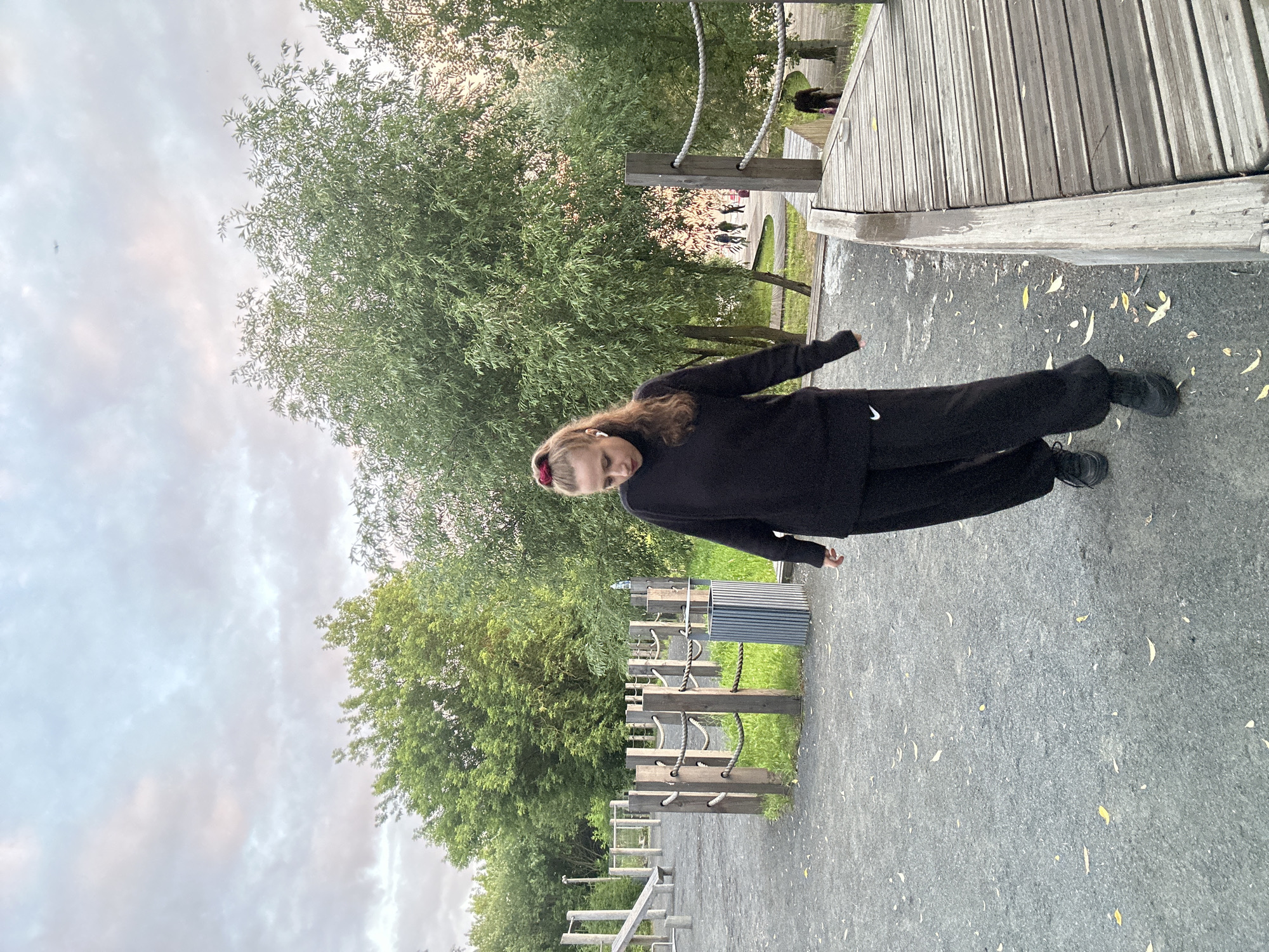 A young person with blonde hair, wearing a black Nike tracksuit, stands on an asphalt path next to a wooden structure. They have a red scrunchie in their hair. The path is scattered with fallen yellow leaves. Behind them, a large green tree with wispy leaves is visible. In the background, there's a wooden walkway and more trees. The sky is overcast with a mix of grey and pink clouds. The scene appears to be an outdoor park or recreational area. The time of day seems to be late afternoon or early evening, given the lighting. The overall mood is casual and relaxed. There is no visible text in the image.