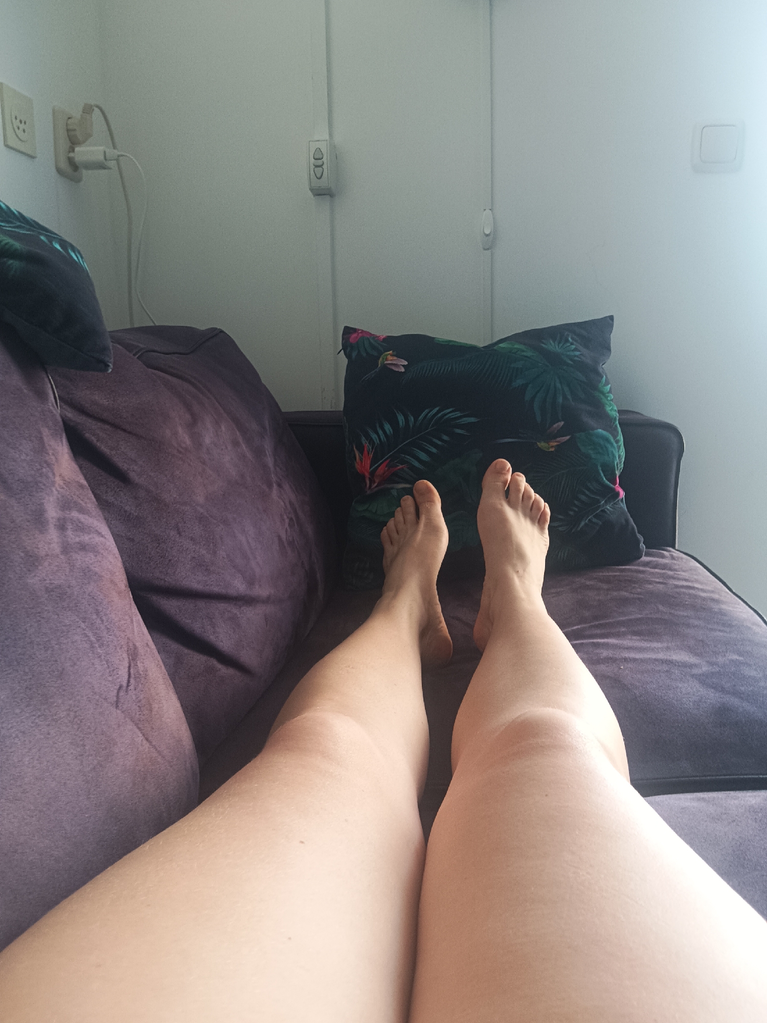 The image displays a person's legs and feet resting on a purple couch. A decorative pillow with a tropical leaf pattern is visible behind the legs. The background consists of a white wall with electrical outlets and a light switch. The scene appears to be indoors, possibly a living room. There is no indication of the city of Makkum, Netherlands, or any specific urban environment.