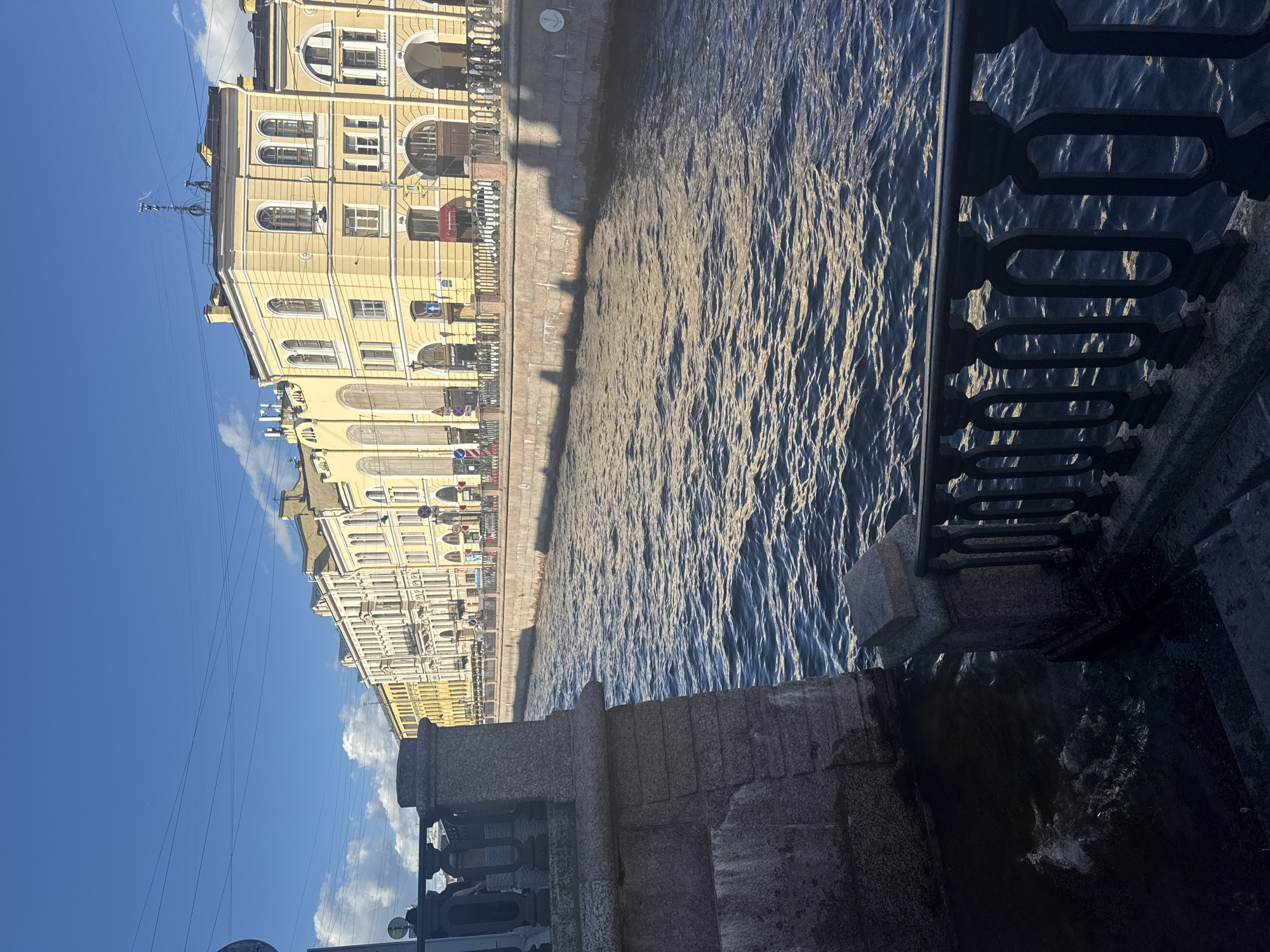 This image captures a view of a canal in Saint Petersburg, Russia, on a bright, sunny day. The water of the canal is choppy, reflecting the sunlight in shimmering patterns. On the left side of the canal, a long, pale yellow building with multiple arched windows and ornate architectural details lines the embankment. Shadows from the building are cast across the water, indicating the sun is high in the sky.
A dark, decorative metal railing runs along the right side of the canal, separating the viewer from the water. Below the railing, there is a stone embankment and steps leading down to the water's edge, some of which are wet. The overall scene suggests a picturesque urban setting, typical of Saint Petersburg's many canals and historic architecture. There are no people visible in the frame, and the focus is on the interplay of light, architecture, and water. The image appears to be taken from a bridge or a higher vantage point, looking down and across the canal.