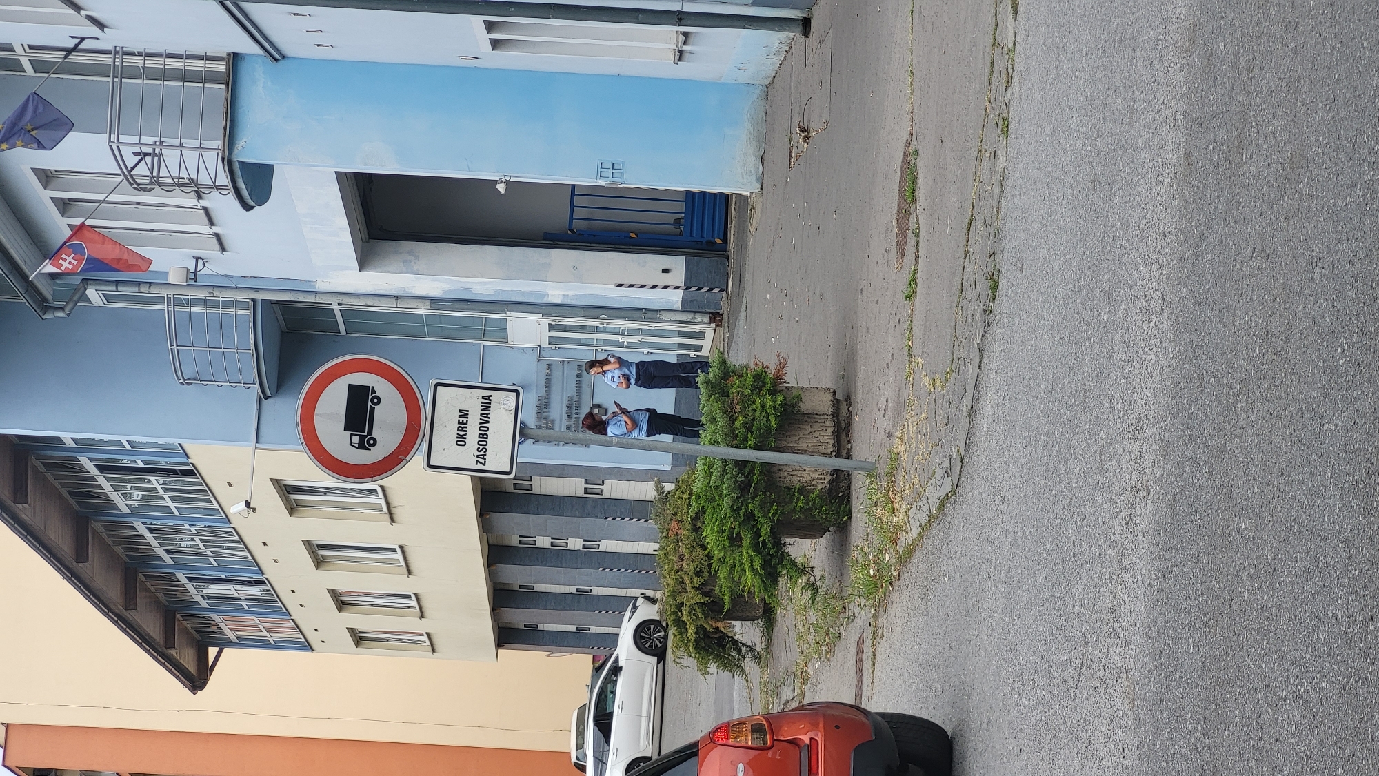 In Prešov, Slovakia, a traffic sign prohibiting trucks is visible in front of a blue building. Below the main sign, a secondary sign reads "OKREM ZÁSOBOVANIA," indicating an exception for supply vehicles. Two women in uniform, possibly security personnel or public servants, are standing near the entrance of the building, facing a window and appearing to be engaged in conversation. One of the women is holding a mobile phone. The scene takes place on a paved area with some greenery in planters. A white car is parked to the left, and the rear of a red car is visible in the foreground, suggesting a street or parking area. The architecture of the building is modern, with multiple windows and a prominent entrance.