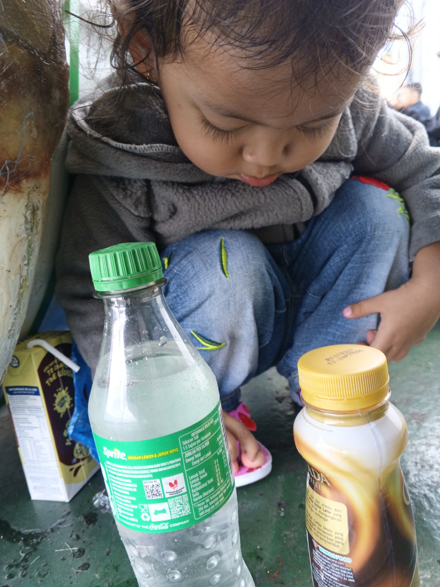 In an outdoor setting, likely a public space in Unknown, Indonesia, a young child is crouching on a surface that appears to be a dirty table or ground. The child, wearing a grey hooded jacket and blue jeans, is looking down with a neutral expression. In the foreground, two bottles are visible. To the left is a clear plastic bottle with a green cap, containing a light-colored liquid, possibly Sprite, as indicated by the label. To the right is a bottle with a yellow cap, containing a brown beverage, possibly coffee. A yellow carton with a straw is positioned behind the Sprite bottle. The scene suggests a casual moment, perhaps during a break or outing.