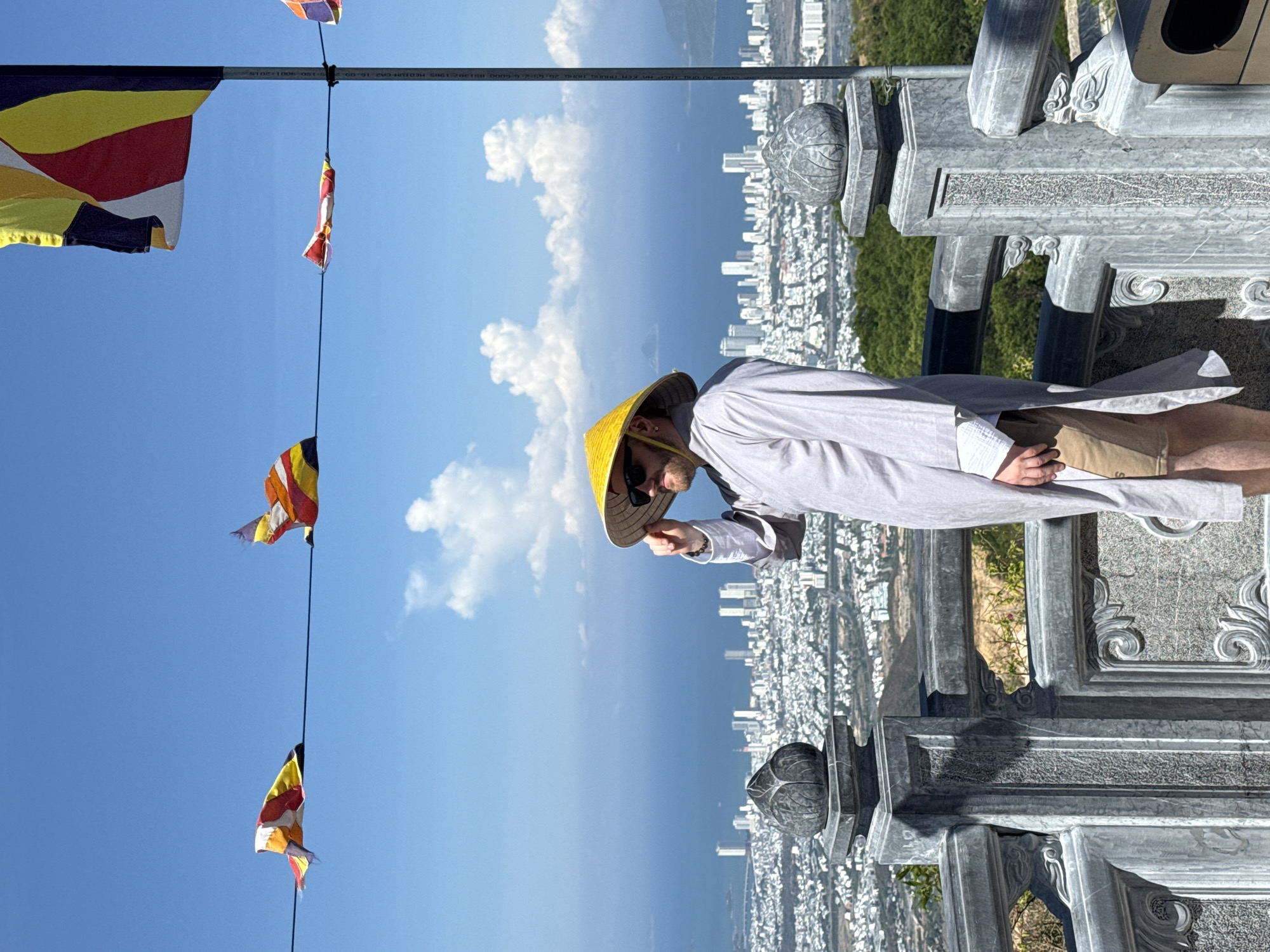 A man wearing a yellow conical hat and sunglasses stands on a stone structure overlooking the city of Nam Nha Trang Ward, Vietnam. He is wearing a light-colored shirt and tan shorts. In the background, there is a view of the city and the sea. A string of Buddhist prayer flags hangs in the air. The sky is blue with scattered clouds.