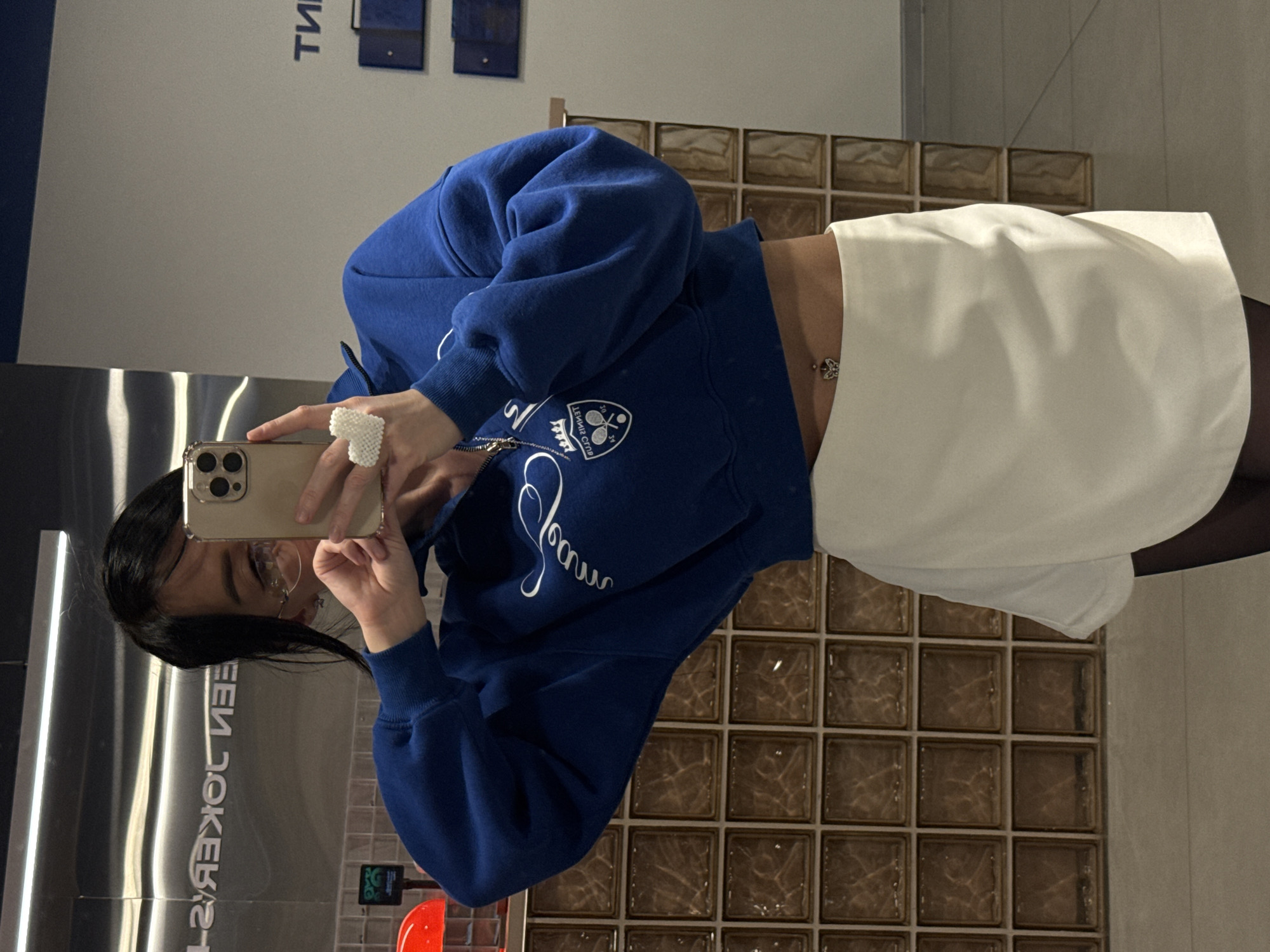 A woman stands indoors, reflected in a shiny, vertical, metallic surface with mirror-like qualities. She is facing the reflective surface and holding a tan-colored smartphone, seemingly taking a selfie or recording a video. She is wearing a royal blue sweatshirt with white writing and a design on the front, coupled with a white skirt and black tights. A bejeweled ring is visible on her finger. Her hair is dark.
Behind her, a wall made of brown, square glass blocks is visible, and further back is a white wall with blue signage on it. Other indiscernible objects are visible on the left side of the frame.
Given the signage appearing on the reflective surface, it's possible that this scene is located in or near a commercial space in Krasnoyarsk, Russia.