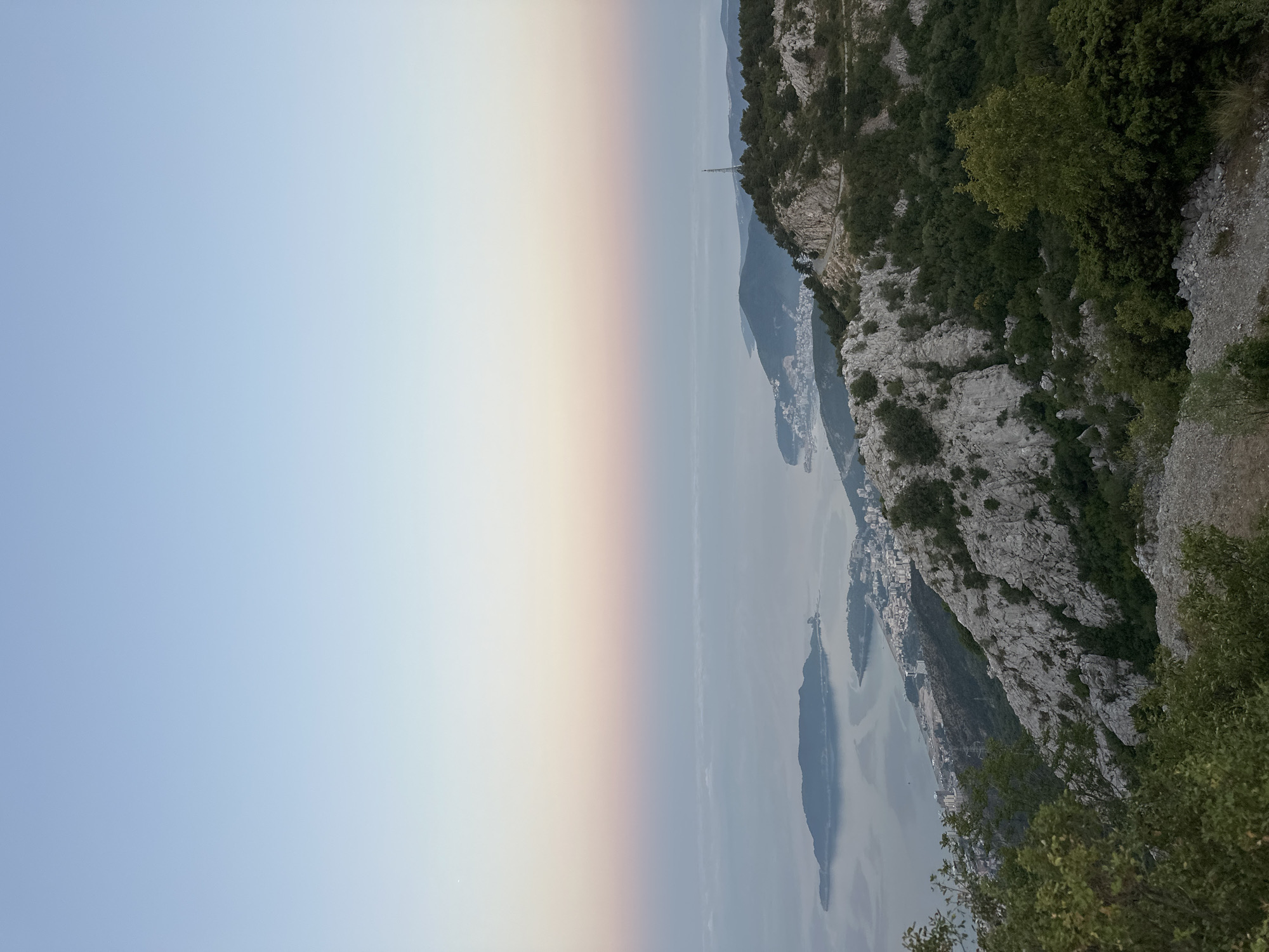 A high-angle, wide shot captures a serene mountainous coastal landscape in Brajici, Montenegro, during twilight. The sky transitions from a pale blue at the zenith to soft hues of pink and orange near the horizon, casting a gentle glow over the scene. Below the sky, a hazy, muted expanse of water stretches out, dotted with distant islands and a coastal town nestled along the shoreline. The town is characterized by a cluster of buildings, some of which appear to be hotels or apartment complexes, indicating a developed area.
In the foreground, rugged, rocky terrain covered in sparse vegetation, including shrubs and small trees, dominates the hillside. A winding dirt road can be seen traversing the mountainous area. The overall impression is one of natural beauty and tranquility, with the human presence subtly integrated into the landscape. No people are visible in the image.