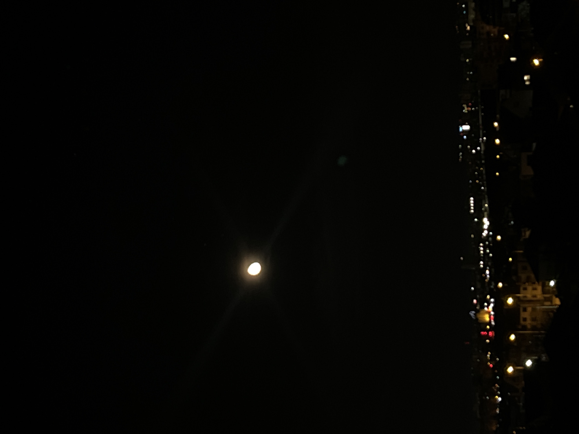 The image shows a night scene in Sofia, Bulgaria. The moon is visible in the sky, and to the right is a blurred view of a city street with buildings and streetlights. The image has a dark and grainy texture.