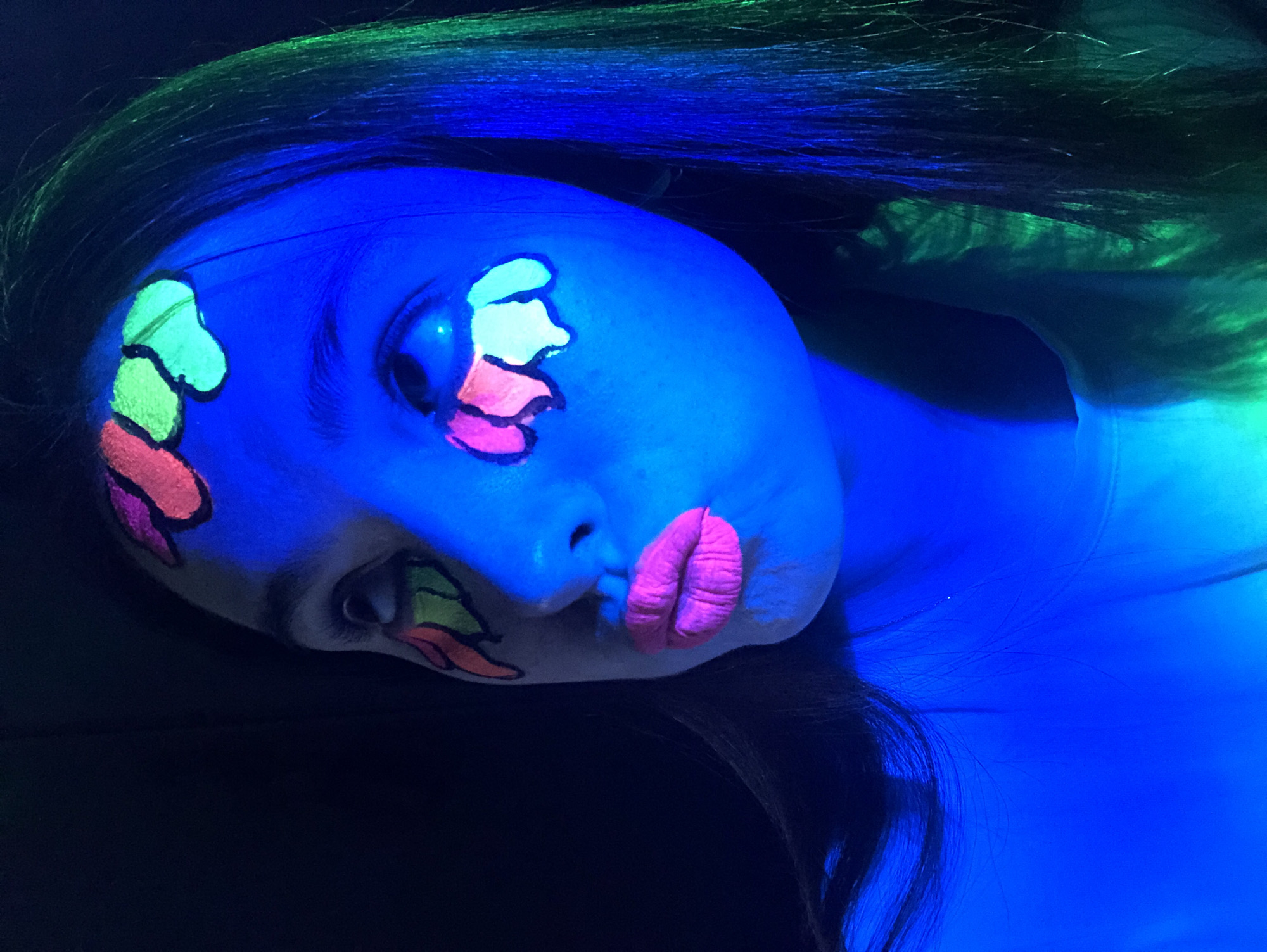 A person with blue-toned skin and face paint lies on a black surface. The face paint is comprised of colorful, abstract shapes near the eyes and mouth. The colors are green, yellow, orange, and pink. The lips are also painted pink. The person's hair is dark and illuminated by green light near the top of the head. The overall lighting effect gives a neon or fluorescent appearance. The location is unknown and not found in the city of Noviy, Russia.