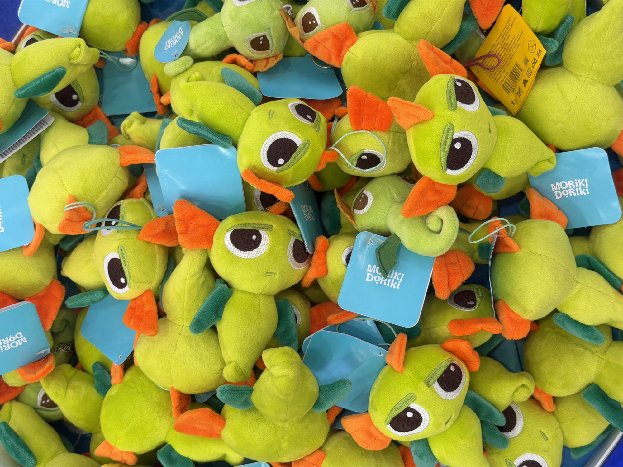 The image shows a close-up view of a pile of plush toys. These toys are light green with orange beaks and green wings. Each toy has a white and brown embroidered eye. Many of the toys have a blue rectangular tag attached, which reads "MORIKI DORIKI." A yellow tag is also visible near the top right of the frame, displaying product information and a barcode. The toys are densely packed together, filling the frame. The image suggests the toys are being sold in a store or market. There is no other information available to verify the location or context.