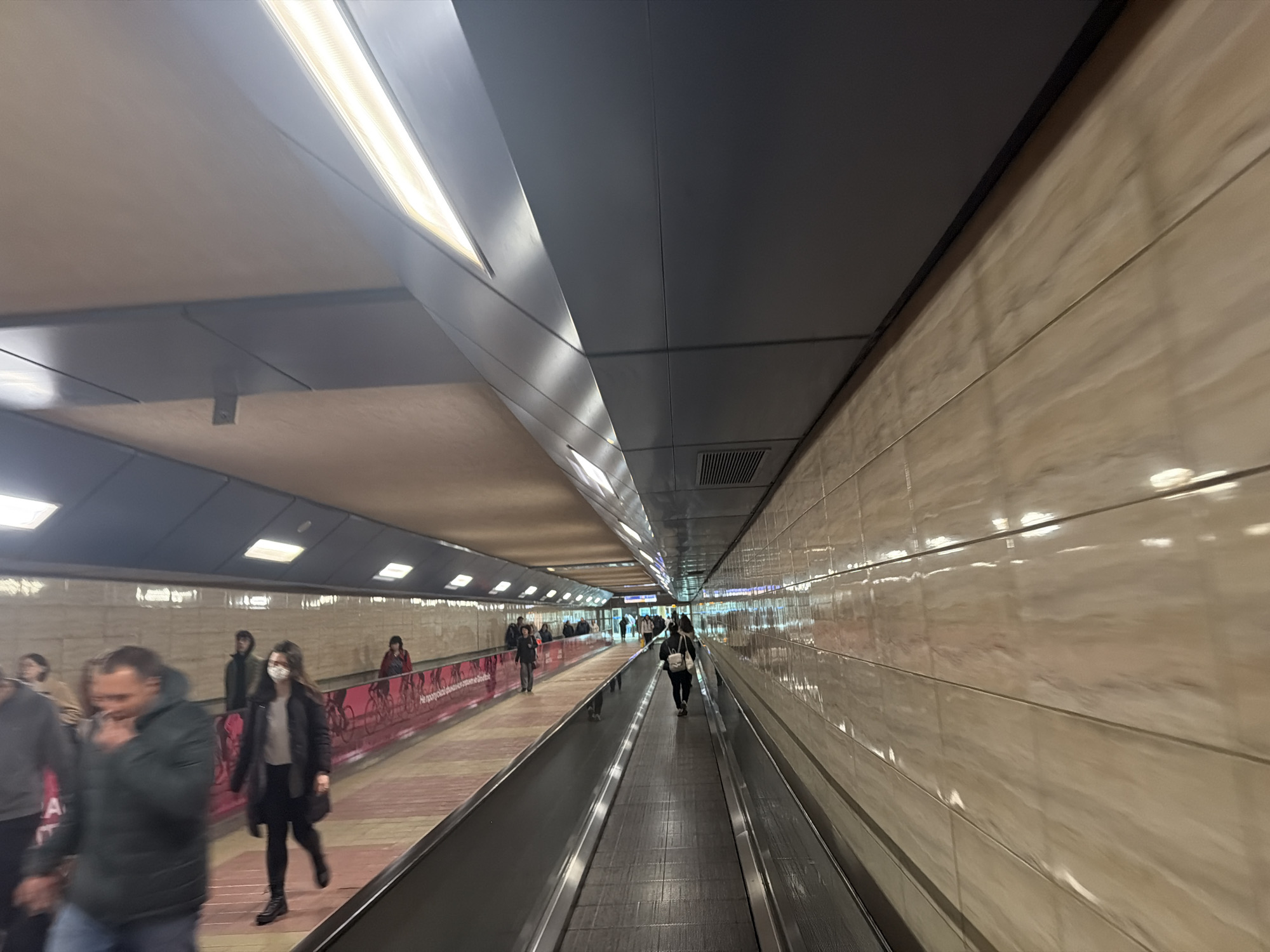 The image shows an indoor pedestrian tunnel with moving walkways. People are walking and standing on the walkways. Some individuals are wearing face masks. The tunnel has a low ceiling with alternating sections of dark gray and light brown. Bright fluorescent lighting is integrated into the ceiling. The walls are covered in horizontal beige tiles. There is a pink and white graphic on the wall near the walkway. Based on visual characteristics, the location appears to be the underground tunnel connecting Terminal 1 and Terminal 2 at Sofia Airport in Sofia, Bulgaria.