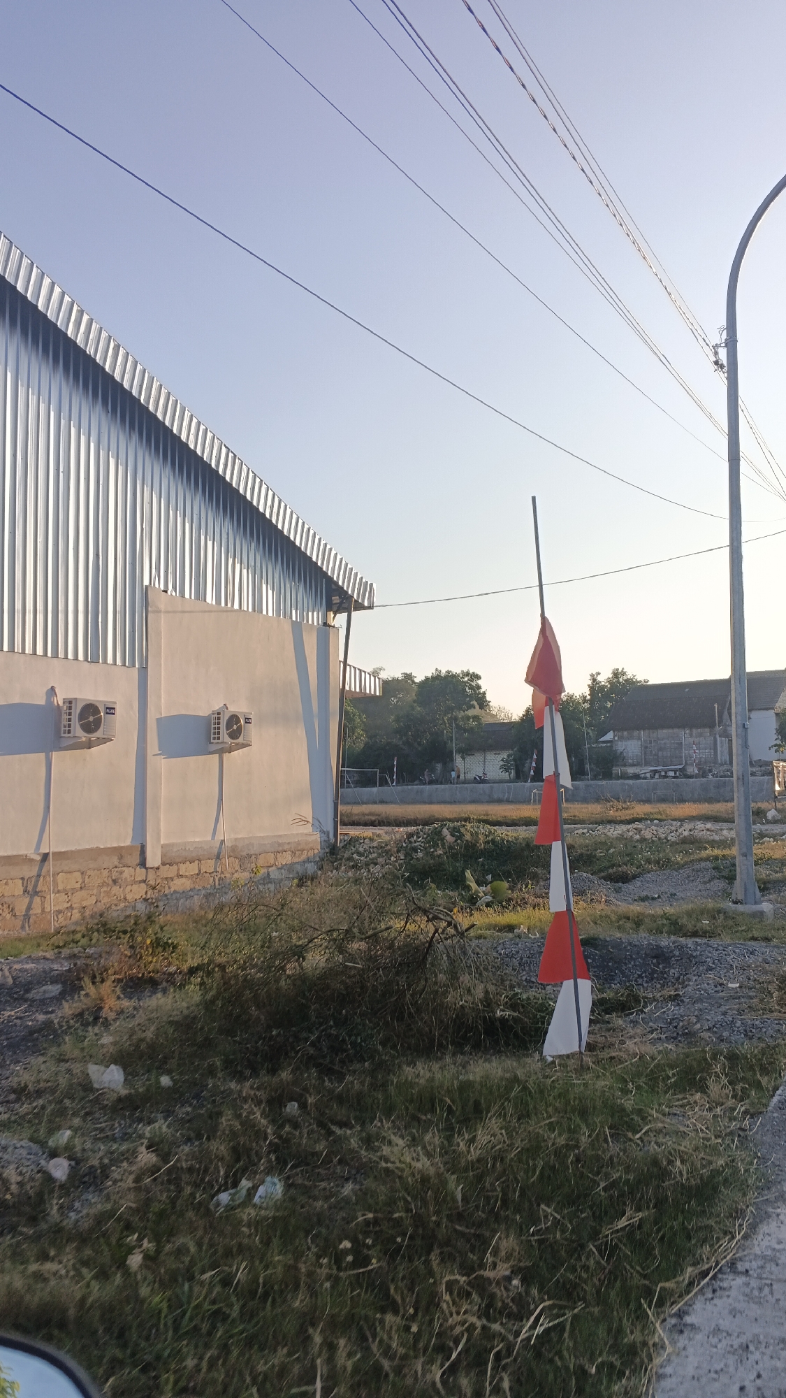 In the city of Soko, Indonesia, a low-angle shot captures a corrugated metal building with a white plastered wall featuring two visible air conditioning units. In the foreground, a pole displays several red and white flags, suggesting a national or ceremonial context. The background reveals a street lamp, overhead power lines, and several residential buildings with tiled roofs. The ground is covered with dry grass, gravel, and some debris. The scene is illuminated by the soft light of either sunrise or sunset, casting long shadows.