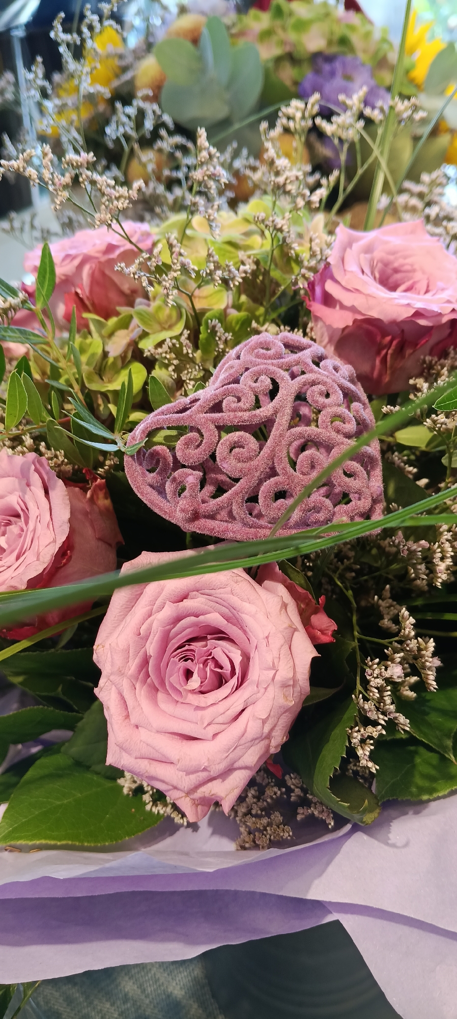 A close-up, eye-level, vertical shot focuses on a bouquet of pink roses and other floral elements. A prominent, ornate pink heart-shaped decoration is placed centrally within the bouquet. The background is a blurred mix of green foliage, smaller white flowers, and hints of yellow and purple blooms. The bouquet is partially wrapped in light purple paper. There is no indication of the location being Linz, Austria.