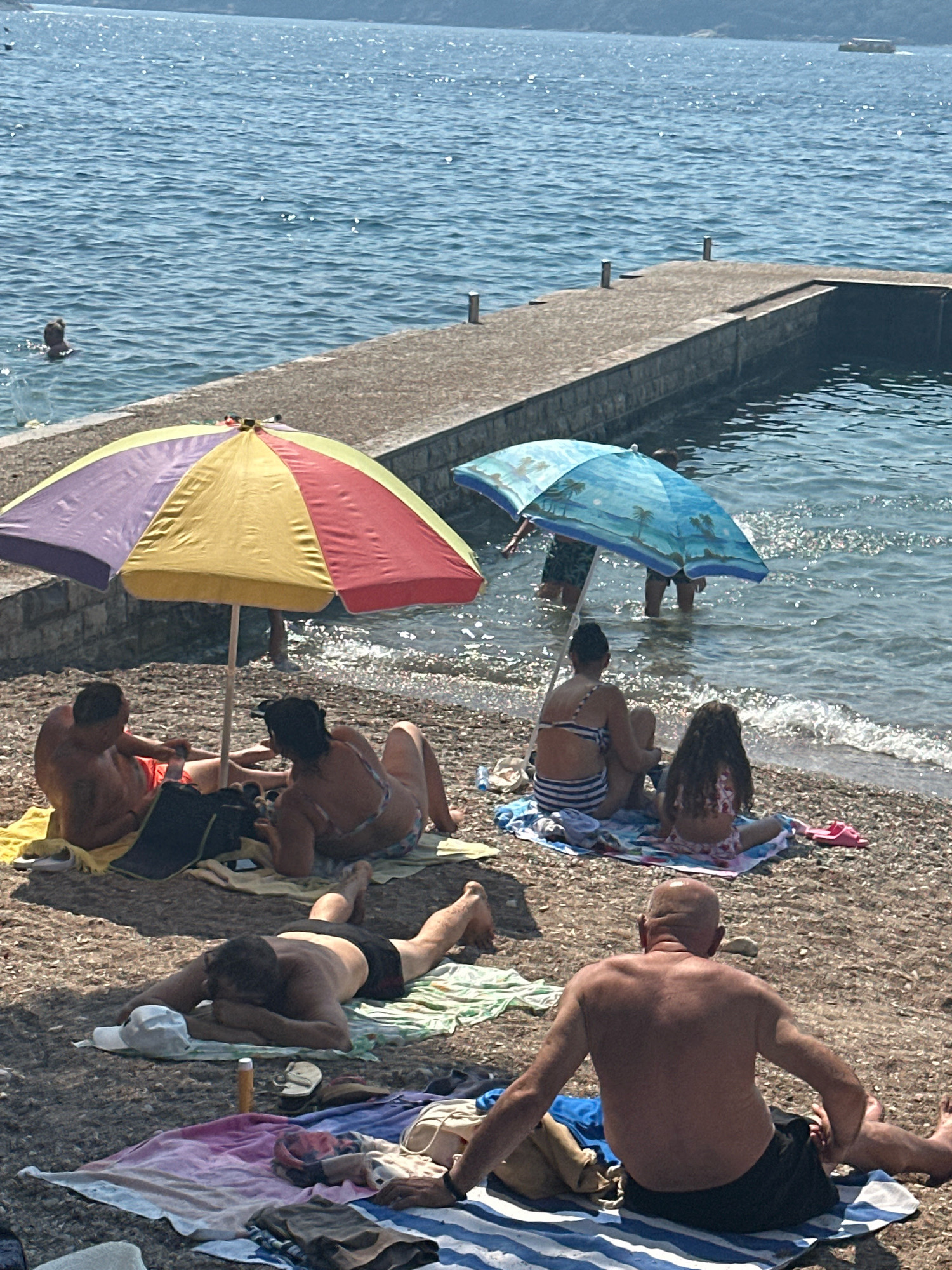 A group of people are relaxing on a pebble beach in Budva, Montenegro, on a sunny day. The water of the Adriatic Sea is clear and blue, with a concrete pier extending into the water. Two beach umbrellas, one multi-colored and the other blue with palm trees, provide shade for some of the beachgoers. Several individuals are lying on towels, some sunbathing and others resting. A few people are wading in the shallow water. There are various beach accessories visible, including towels, bags, flip-flops, and sunscreen. In the background, a boat can be seen on the horizon.