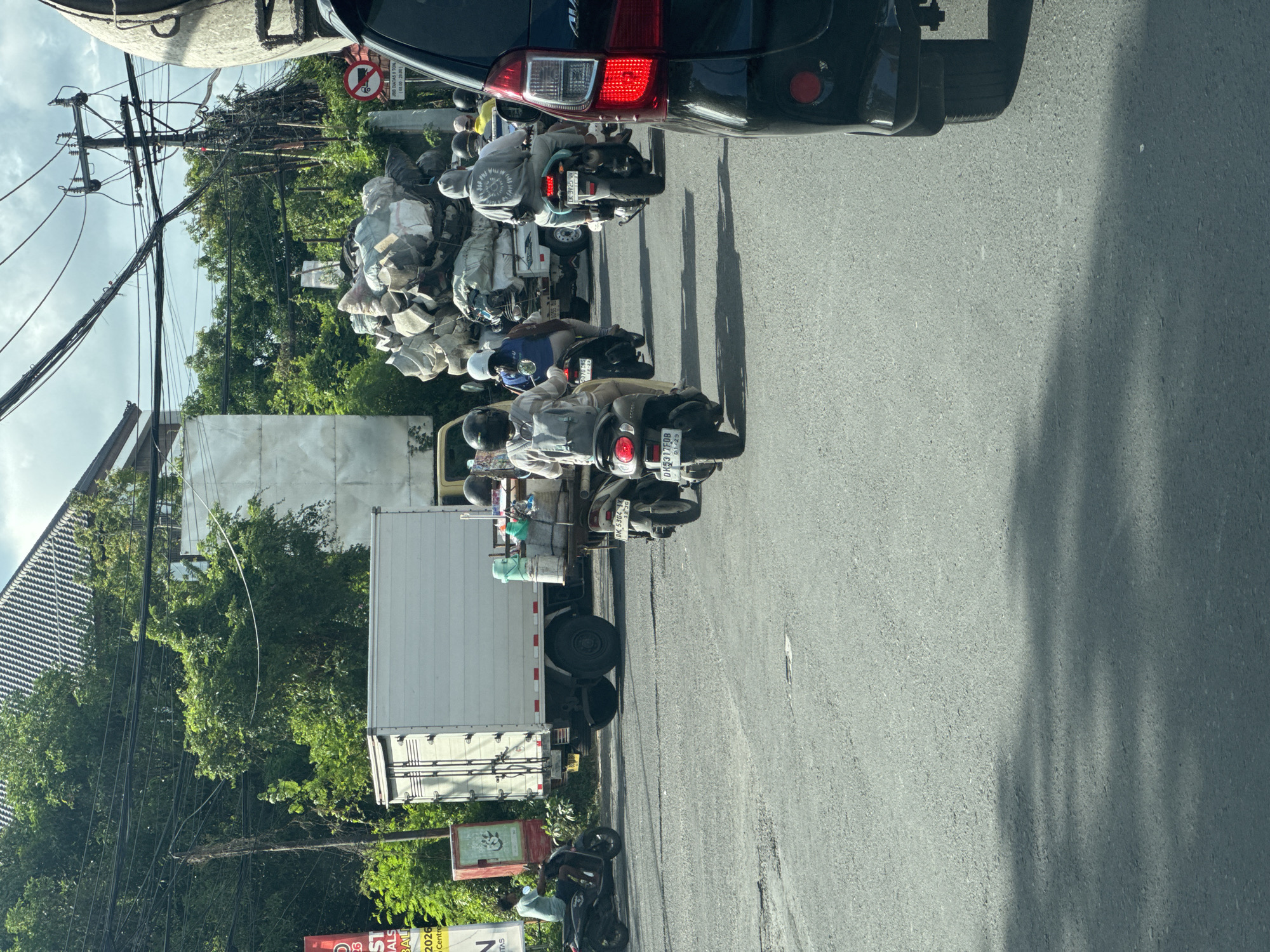 The image captures a congested street scene in Kuta Selatan, Indonesia, characterized by a traffic jam involving cars, motorcycles, and a small truck. Several motorcycles are heavily loaded with bulky materials, possibly recyclables, suggesting a transportation method for goods or waste. The scene is framed from inside a vehicle, with a portion of the car's rear visible. Power lines are strung overhead, and commercial signage indicates a local business. The overall atmosphere suggests a busy urban environment in a developing country.