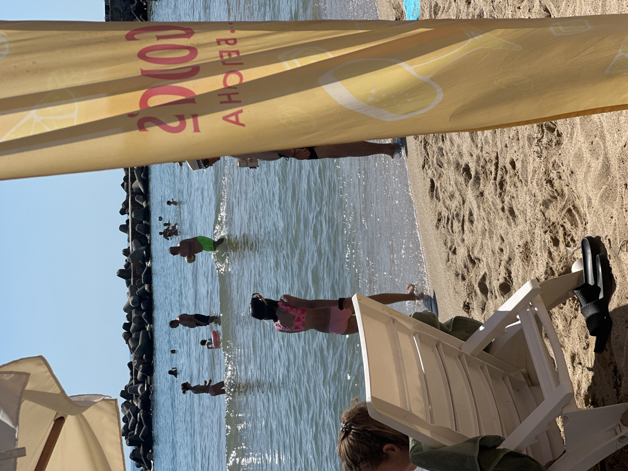 The image depicts a beach scene on a sunny day. In the foreground, a partially visible person is seated in a white lounge chair on the sand, with a green towel draped over it. Behind the chair, a yellow banner with red lettering hangs, obscuring part of the background. The banner displays an image of lemons and text. The middle ground shows a breakwater made of dark, concrete tetrapods, separating the sandy beach from the ocean. Several people are visible in the shallow water, some standing, and one appears to be wading. The water is clear and reflects the sunlight, with small waves lapping at the shore. The sky above is a clear blue. The overall atmosphere suggests a leisurely day at the beach. Visible text on the banner reads "COVER" and "AHOREA".